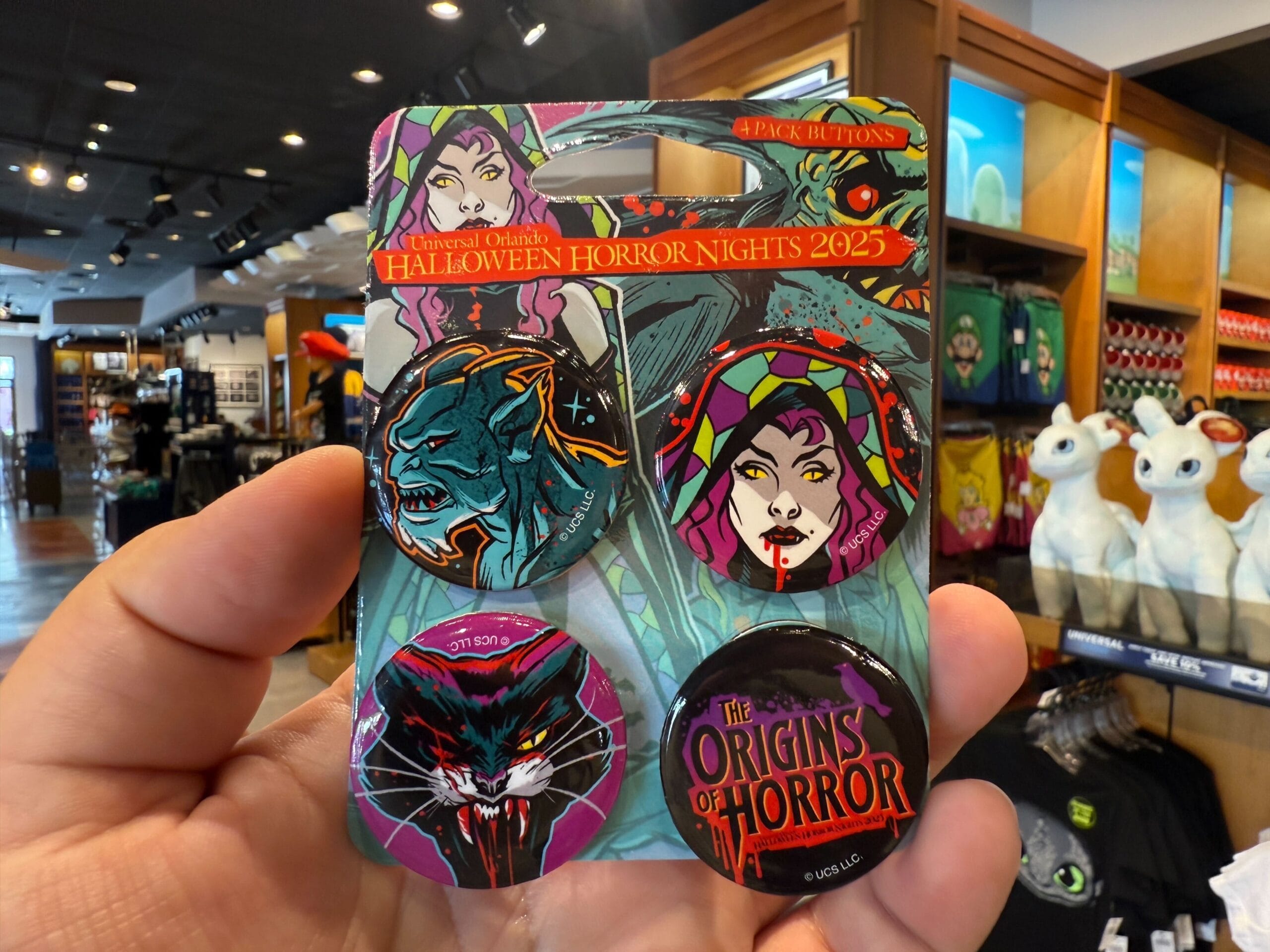 A hand holds four Universal Orlando Resort Halloween Horror Nights 2025 merchandise buttons with monster and vampire designs.