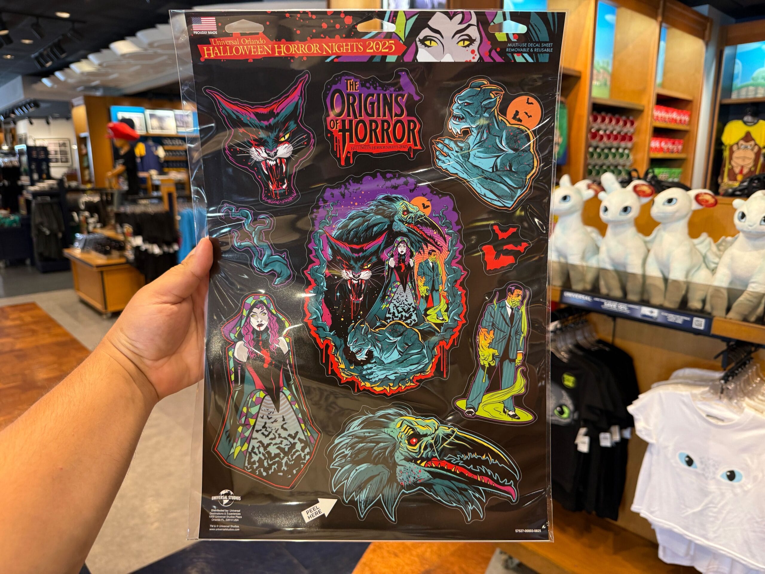 A hand holds a "The Origins of Horror" Halloween sticker sheet among HHN merch at Universal Orlando Resort.