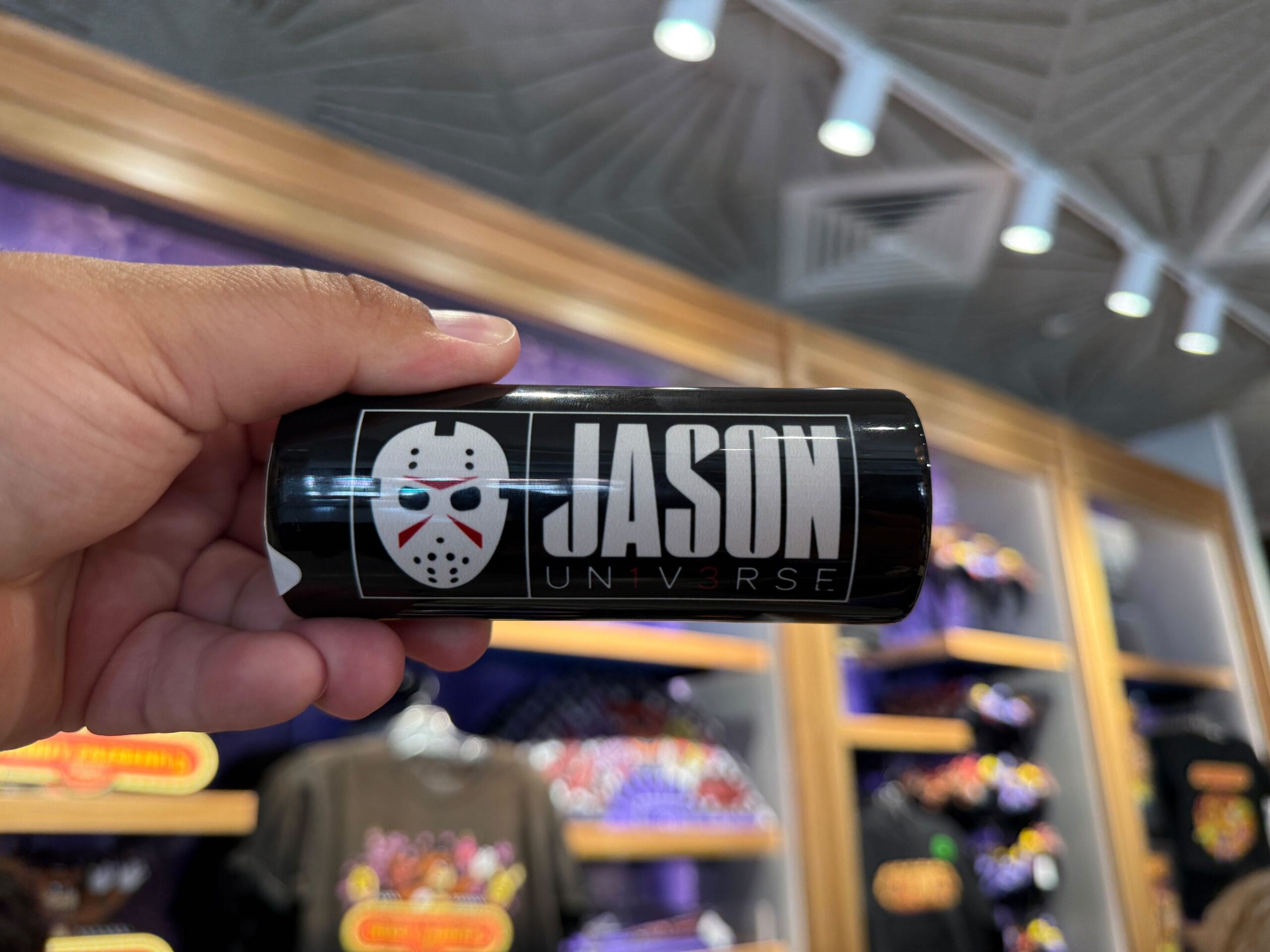 A hand holds HHN Merch with a hockey mask and "JASON UN1VERSE" at Universal Orlando Resort, shirts in the background.