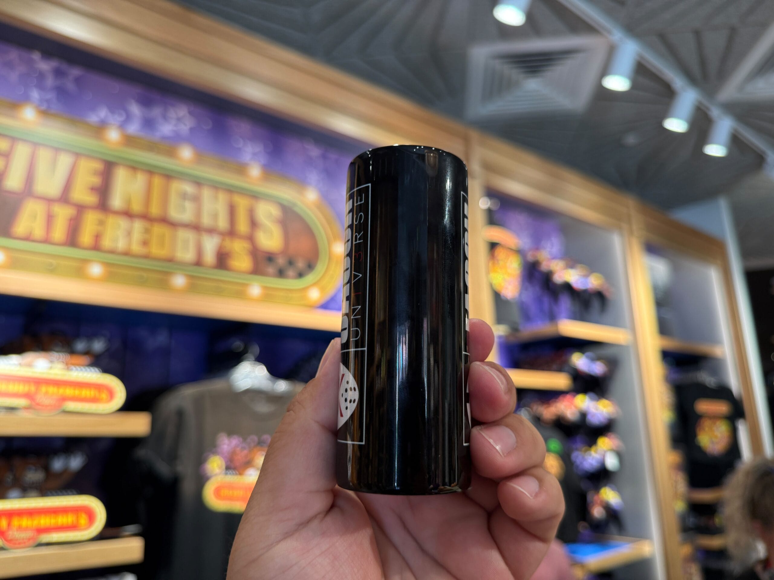A hand holds black HHN Merch in front of Five Nights at Freddy's items at Universal Orlando Resort.