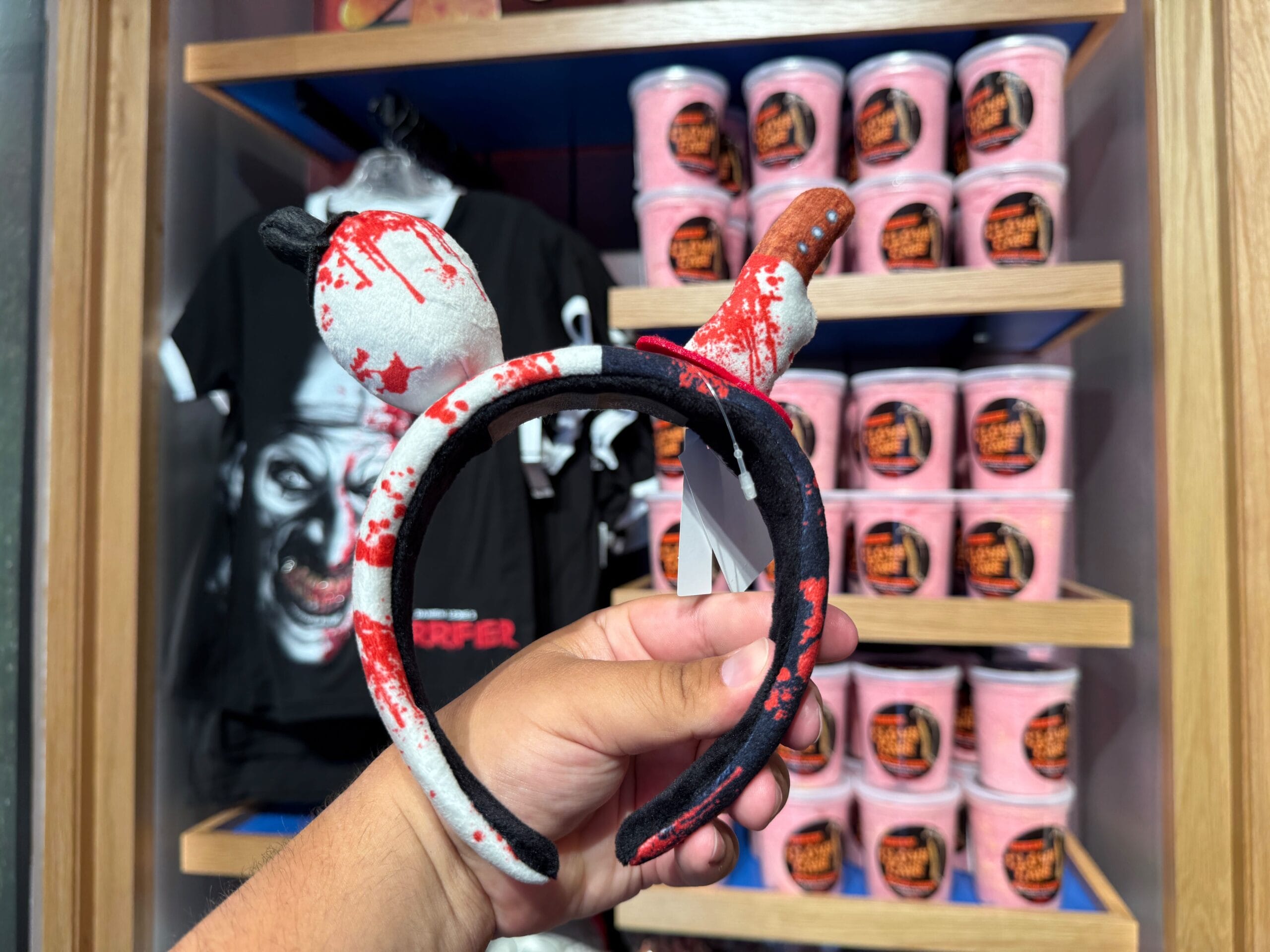 A hand holds bloody knife-themed Mickey ears headband amid HHN merch at Universal Orlando Resort.