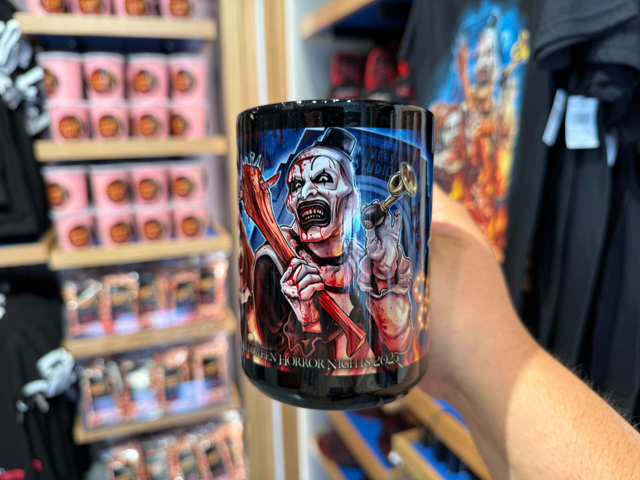 A hand holds a black mug featuring a scary clown at Universal Orlando Resort, surrounded by HHN Merch on shelves.