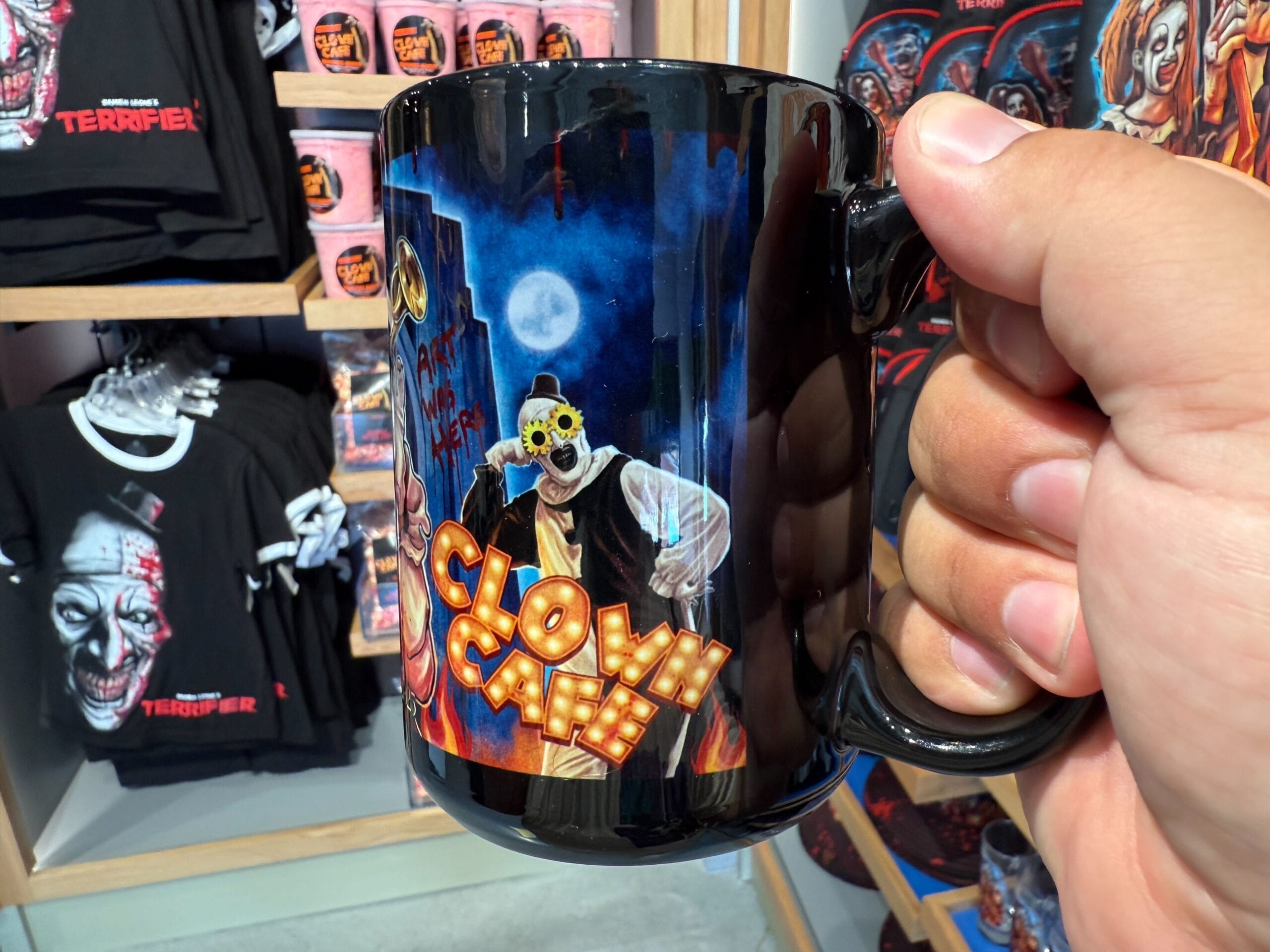 A hand holds a mug with a sinister clown and "Clown Café"—perfect HHN Merch at Universal Orlando Resort.