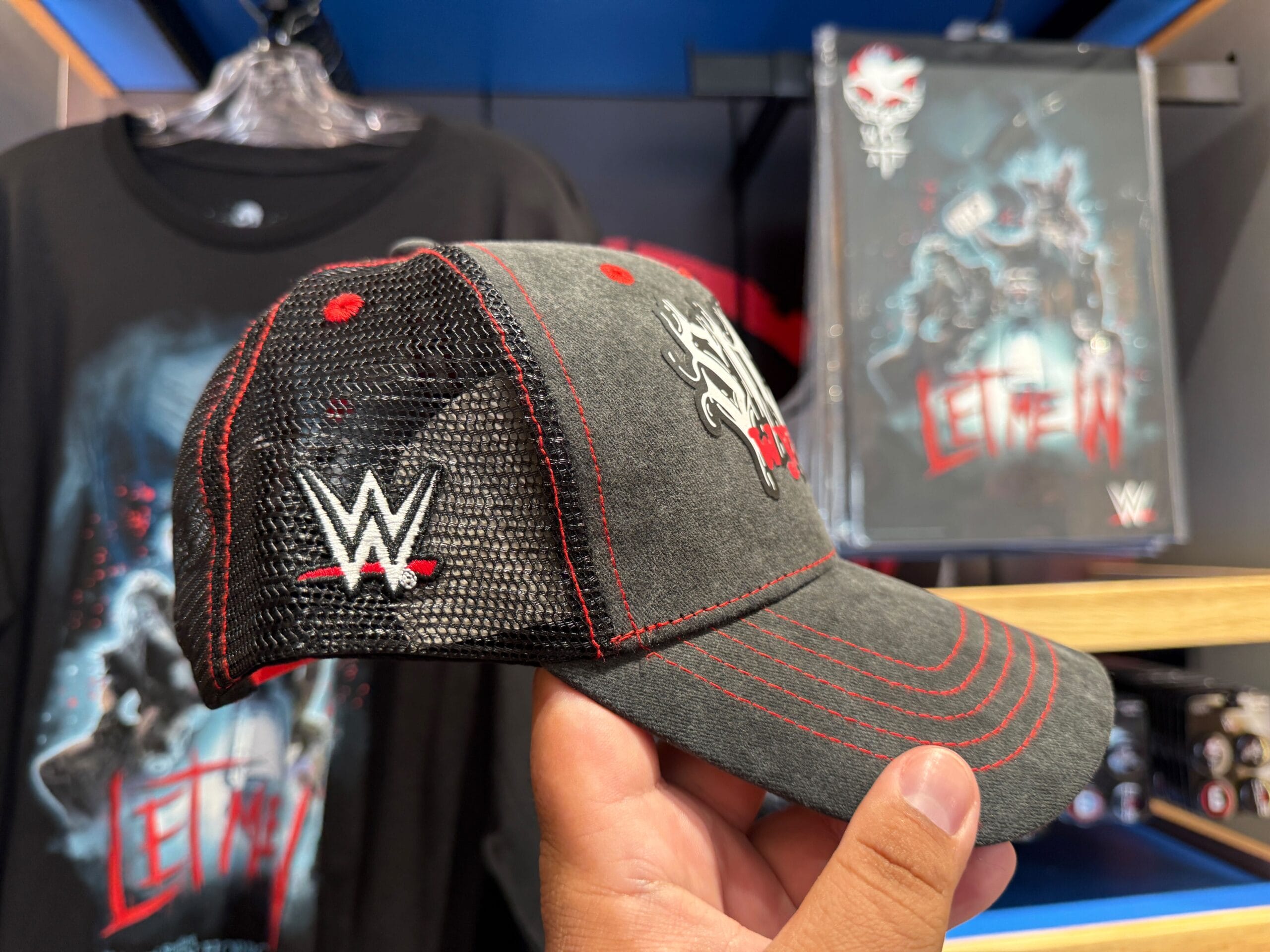 A hand holds a WWE cap in front of "Let Me In" themed HHN merch on display at Universal Orlando Resort.
