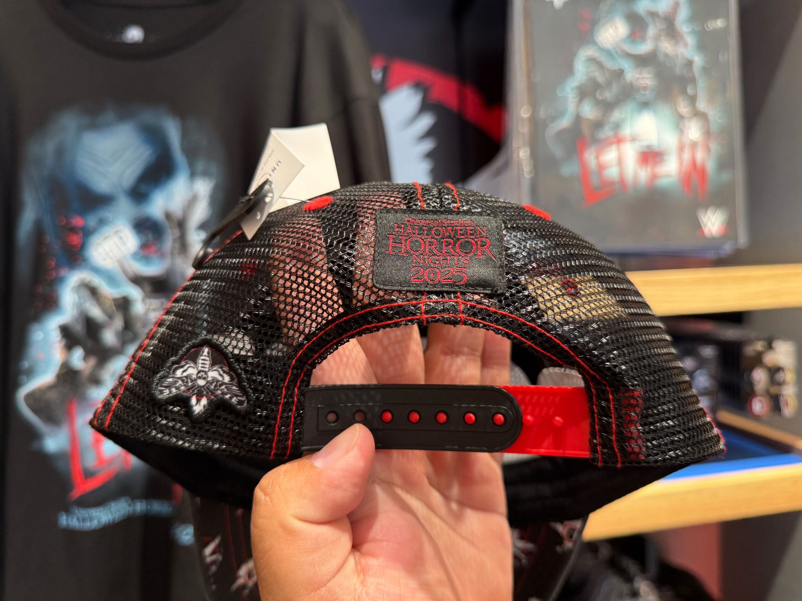 A hand holds a black mesh cap reading "Halloween Horror Nights 2025" at Universal Orlando Resort, merch in the background.
