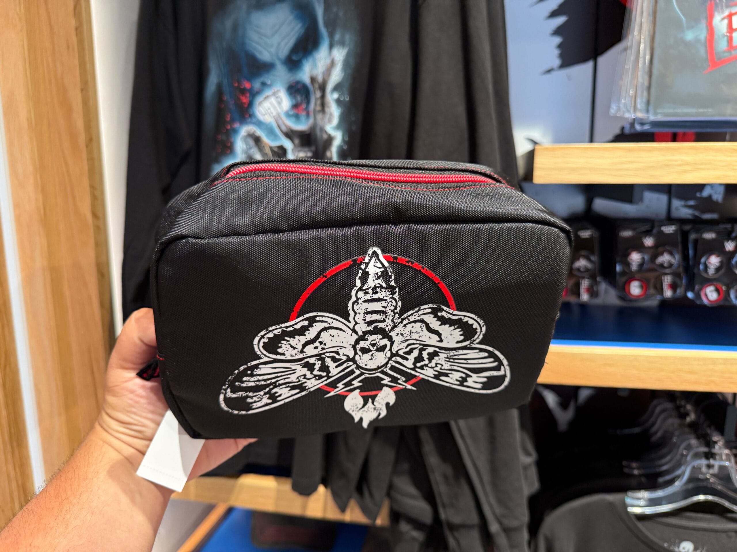 A hand holds a black pouch with a white and red moth, part of exclusive HHN Merch in front of T-shirts on display.