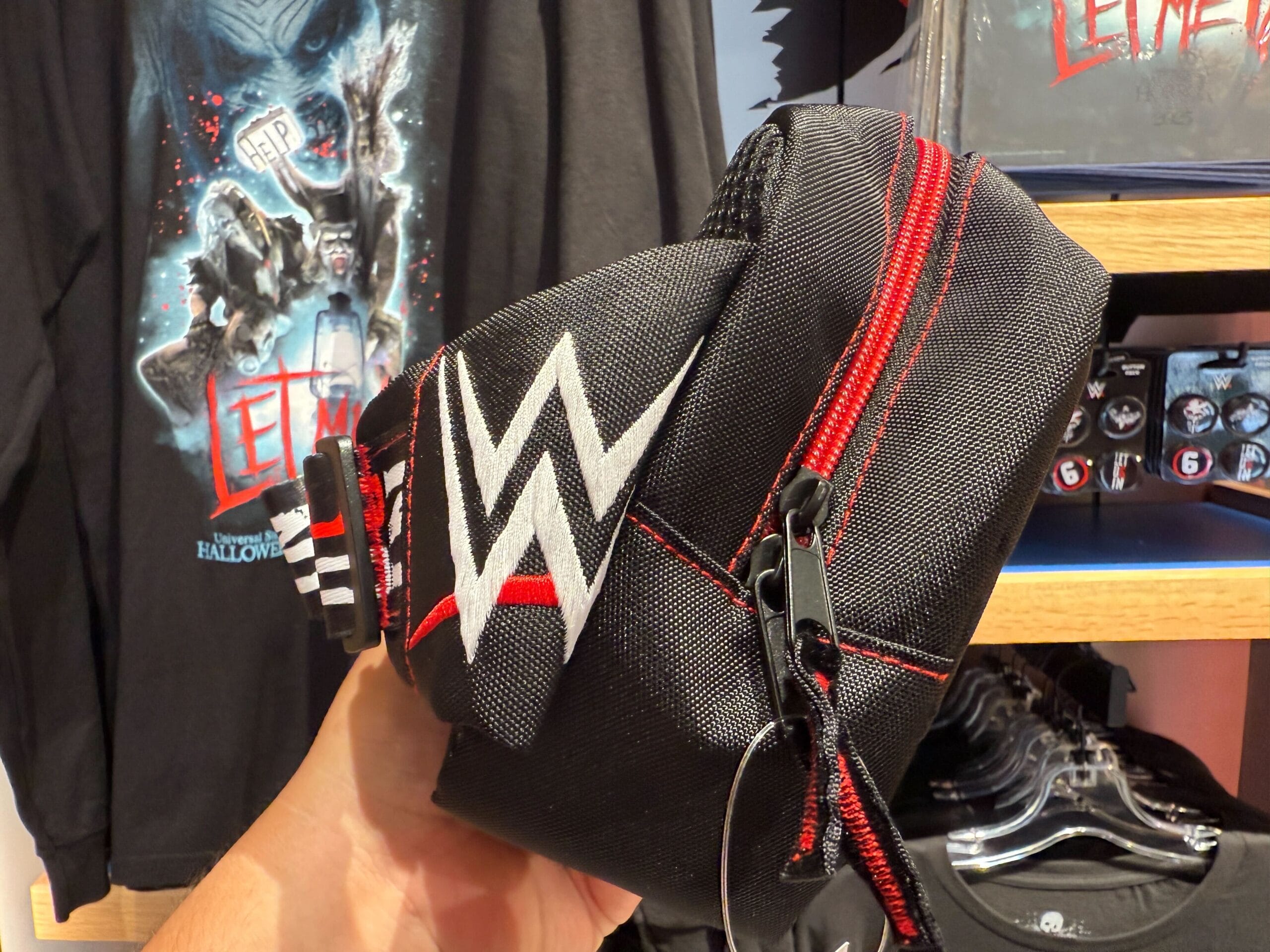 A hand holds a black WWE waist bag with a red zipper, surrounded by HHN merch at Universal Orlando Resort.