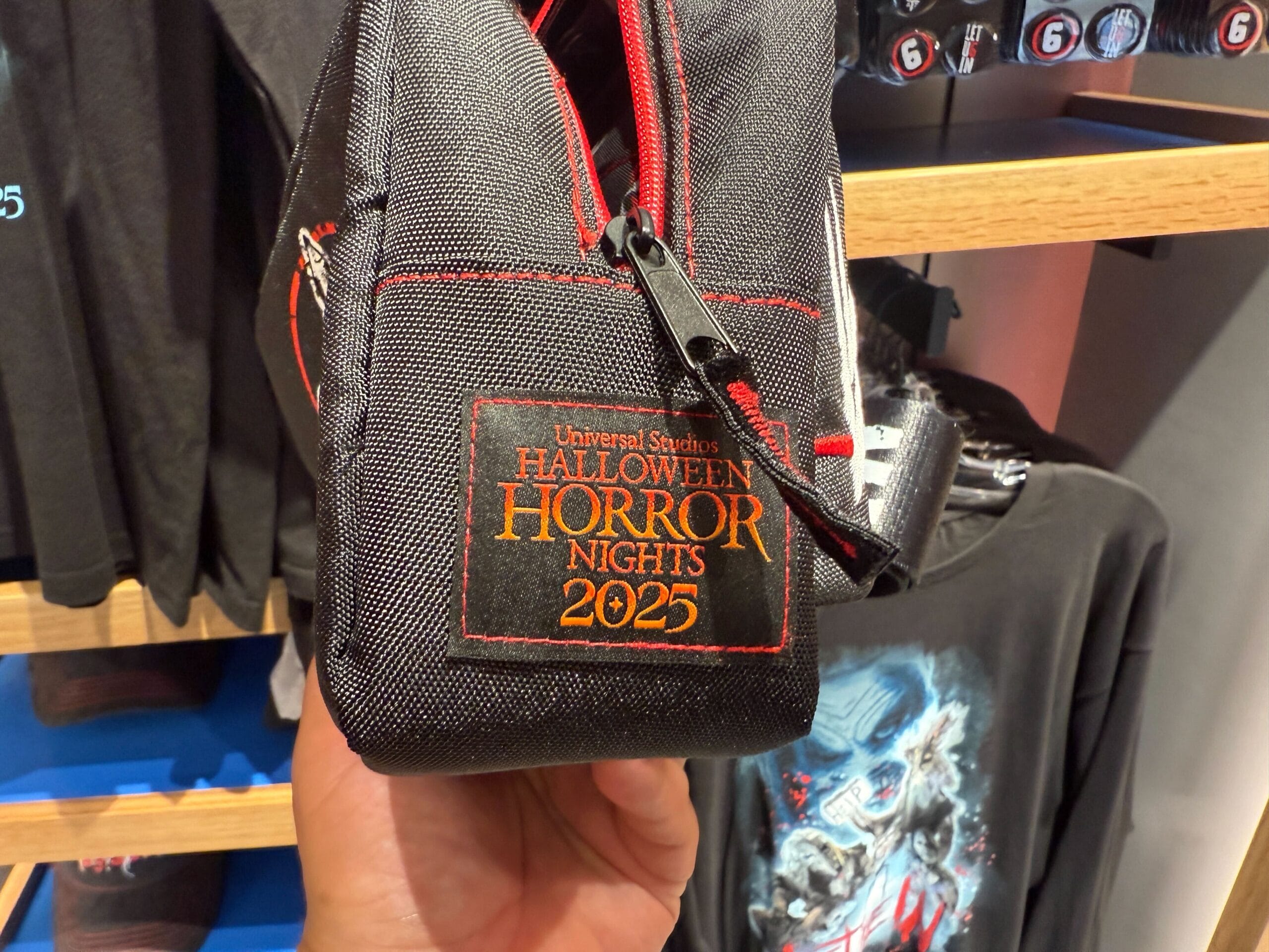 A hand holds Universal Orlando Resort's HHN Merch—a black pouch with "Halloween Horror Nights 2025" embroidered in orange and yellow.