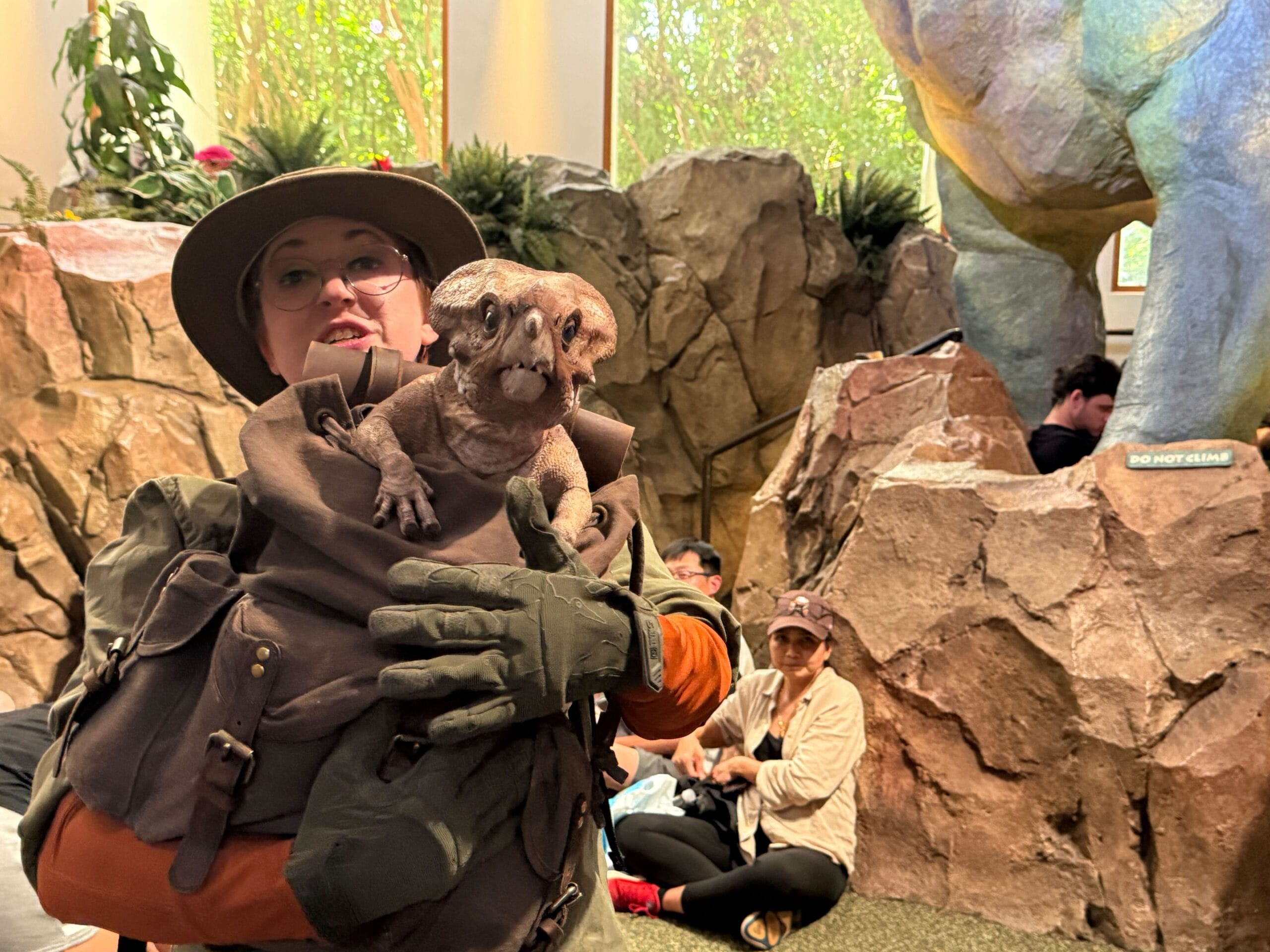 A person in safari attire holds a small dinosaur puppet indoors; people and rocks are in the background.