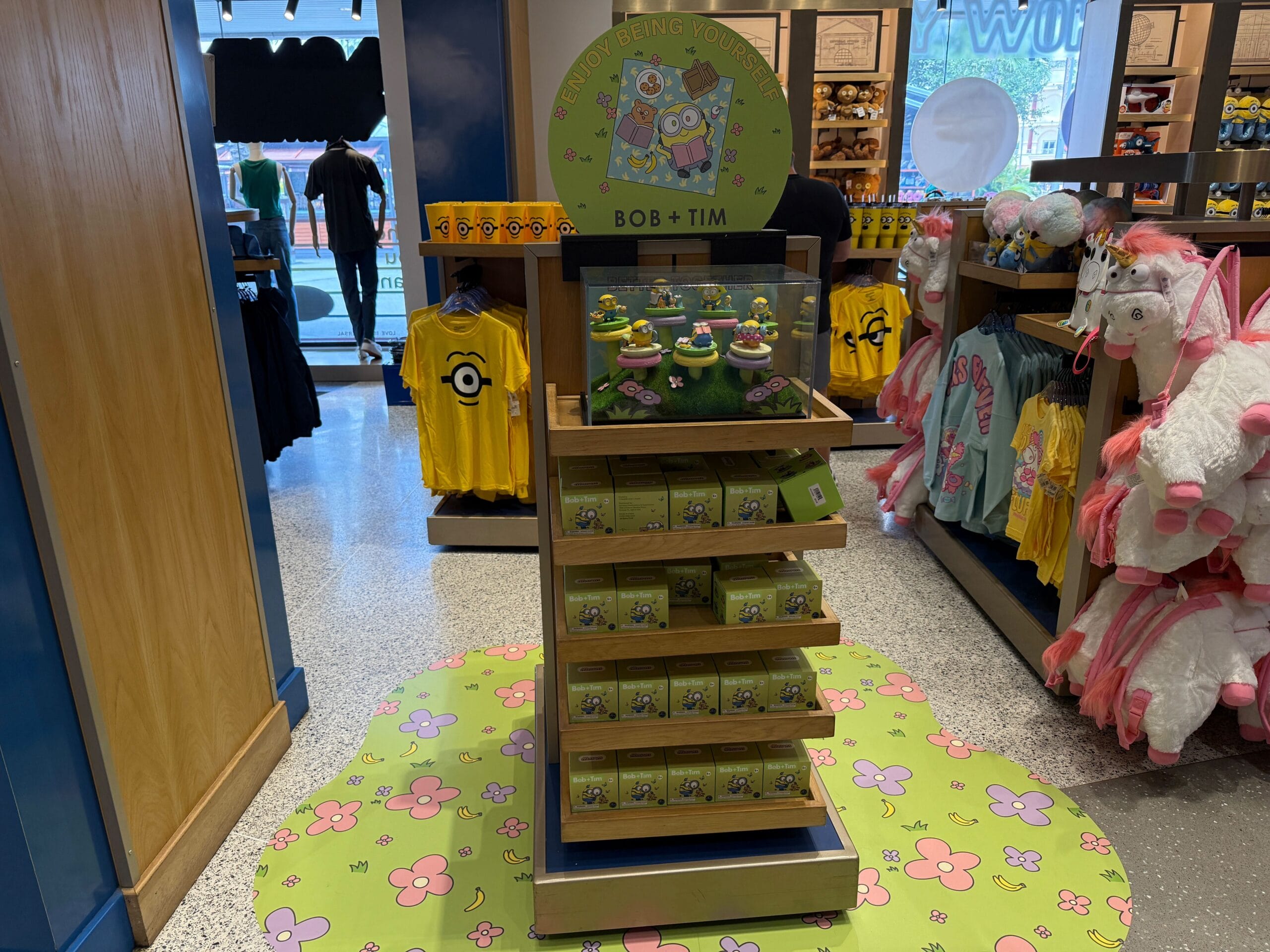 Minions-themed store display with Bob and Tim mystery figure boxes, surrounded by colorful shirts and unicorn plush toys.
