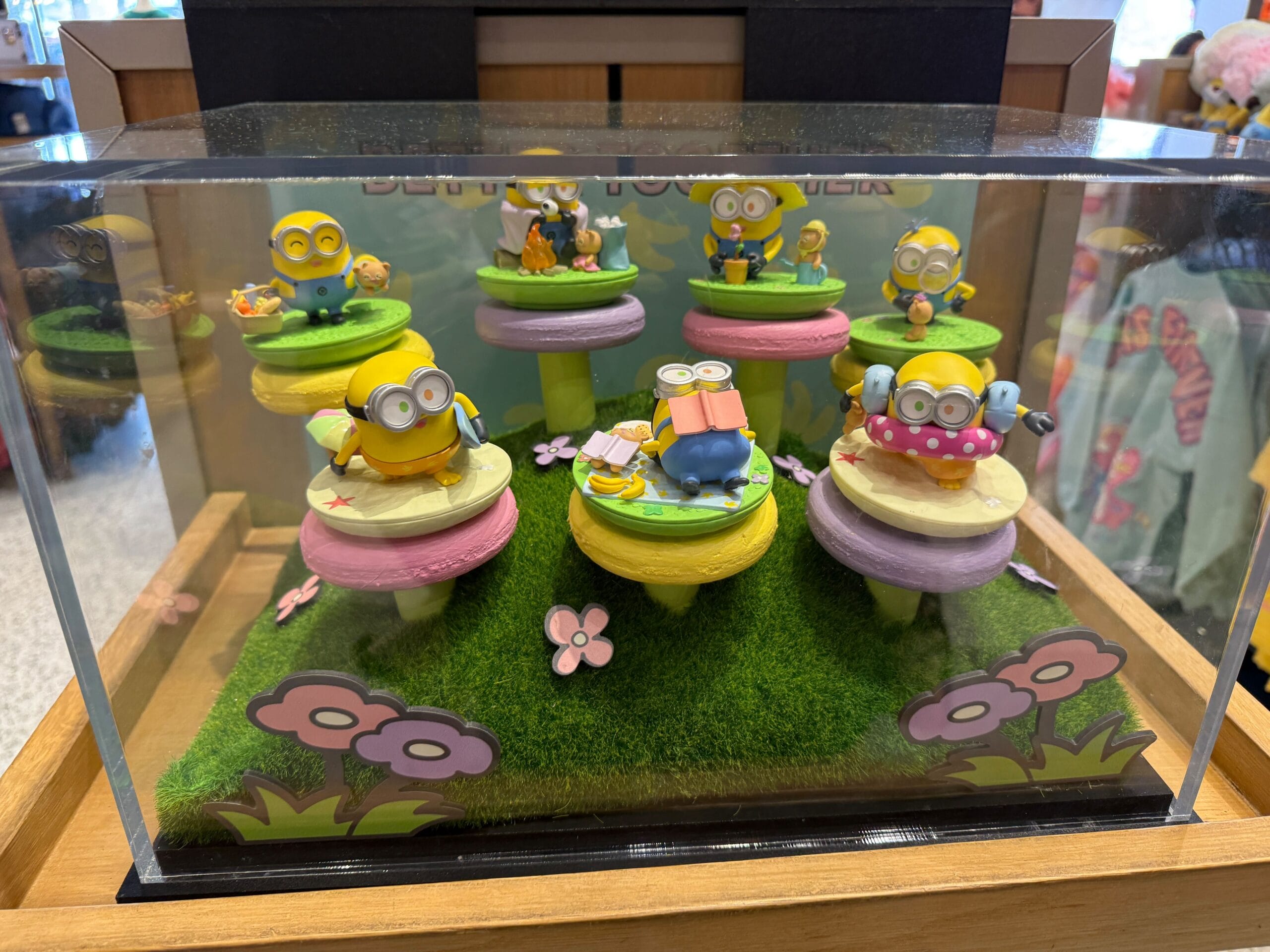 Miniature Bob and Tim Minion figurines displayed on colorful pedestals inside a glass case decorated with flowers and grass.