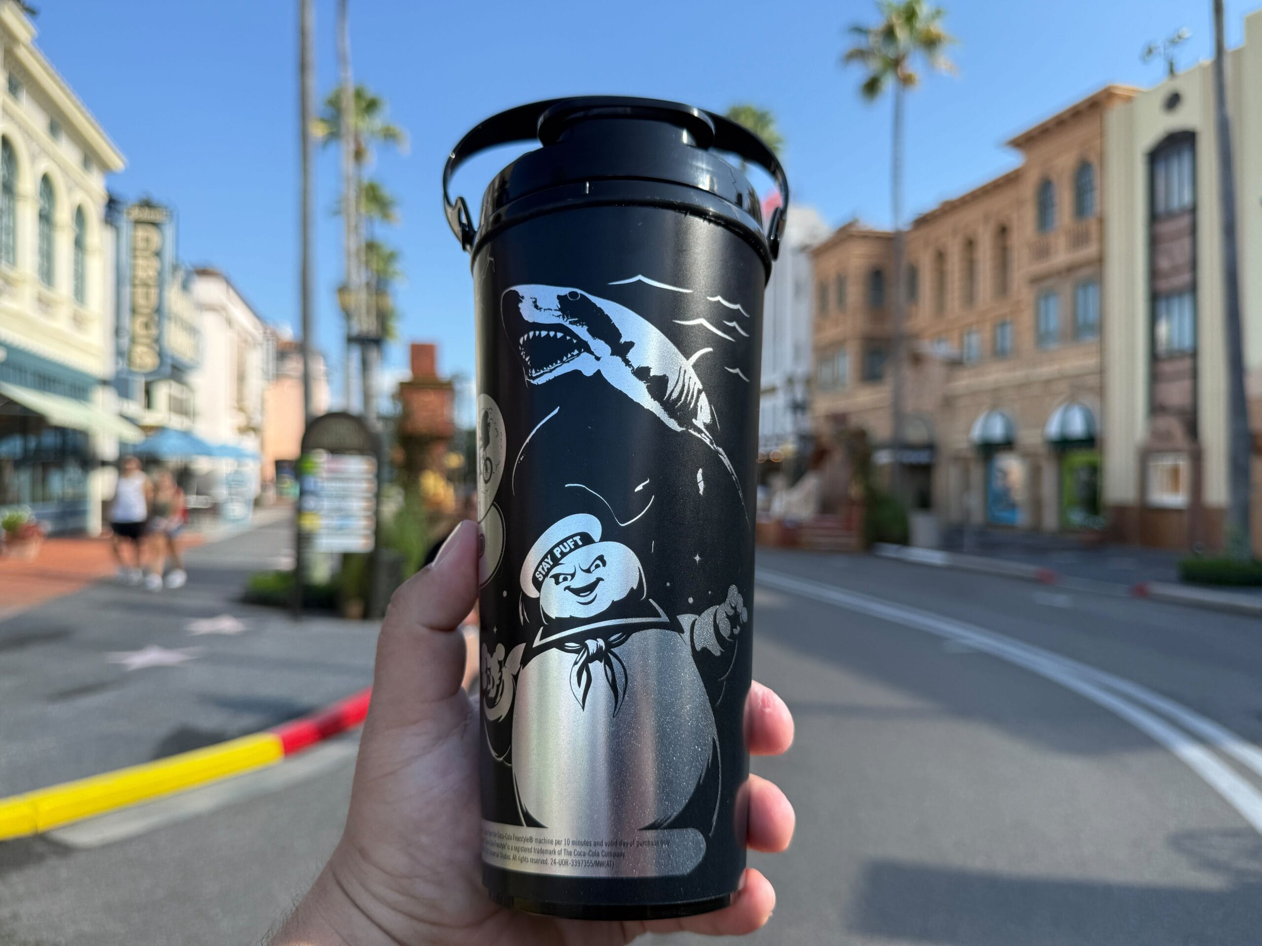 A hand holds a black UOAP Coca-Cola Freestyle Cup with Stay Puft and a shark, buildings and palm trees in the background.