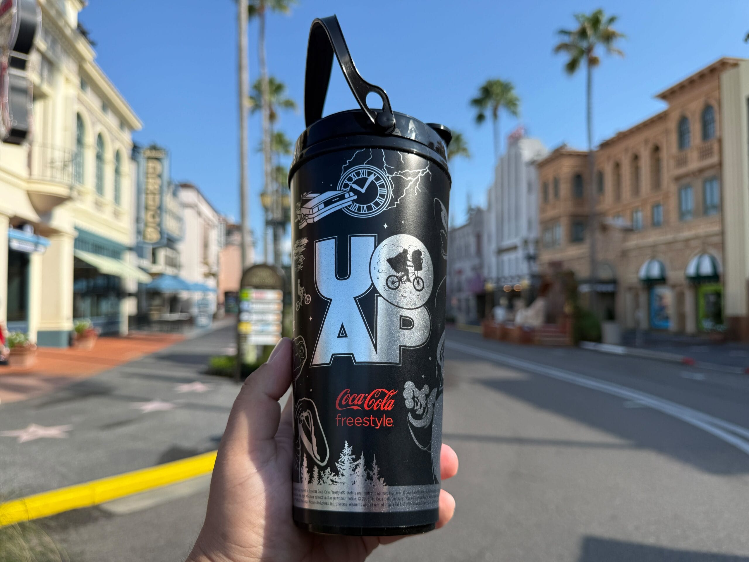 A hand holds a black Coca-Cola Freestyle Cup during UOAP Passholder Appreciation Days on a sunny, palm-lined street.
