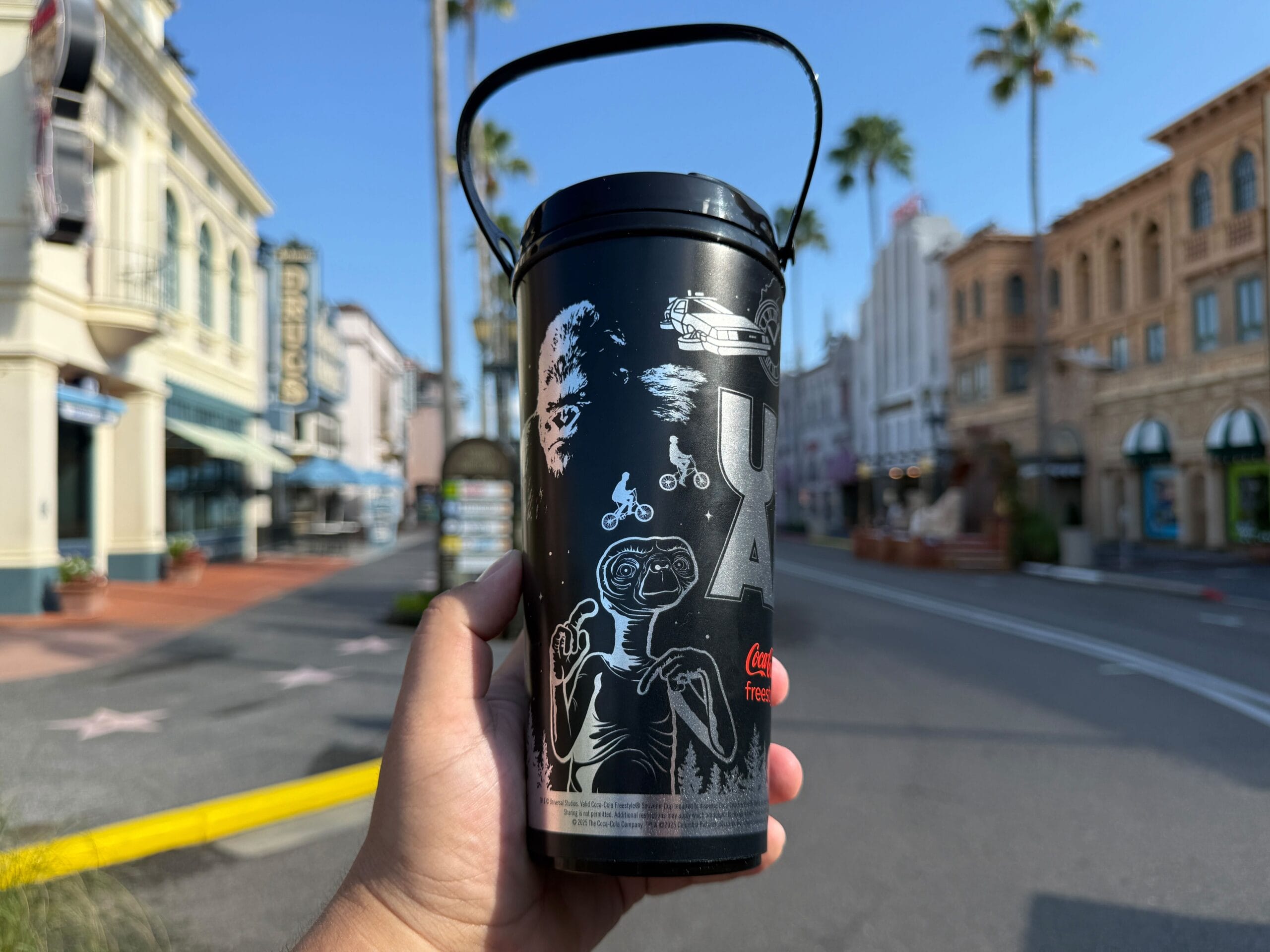 A hand holds a black UOAP Coca-Cola Freestyle Cup on an empty street lined with palm trees and buildings.
