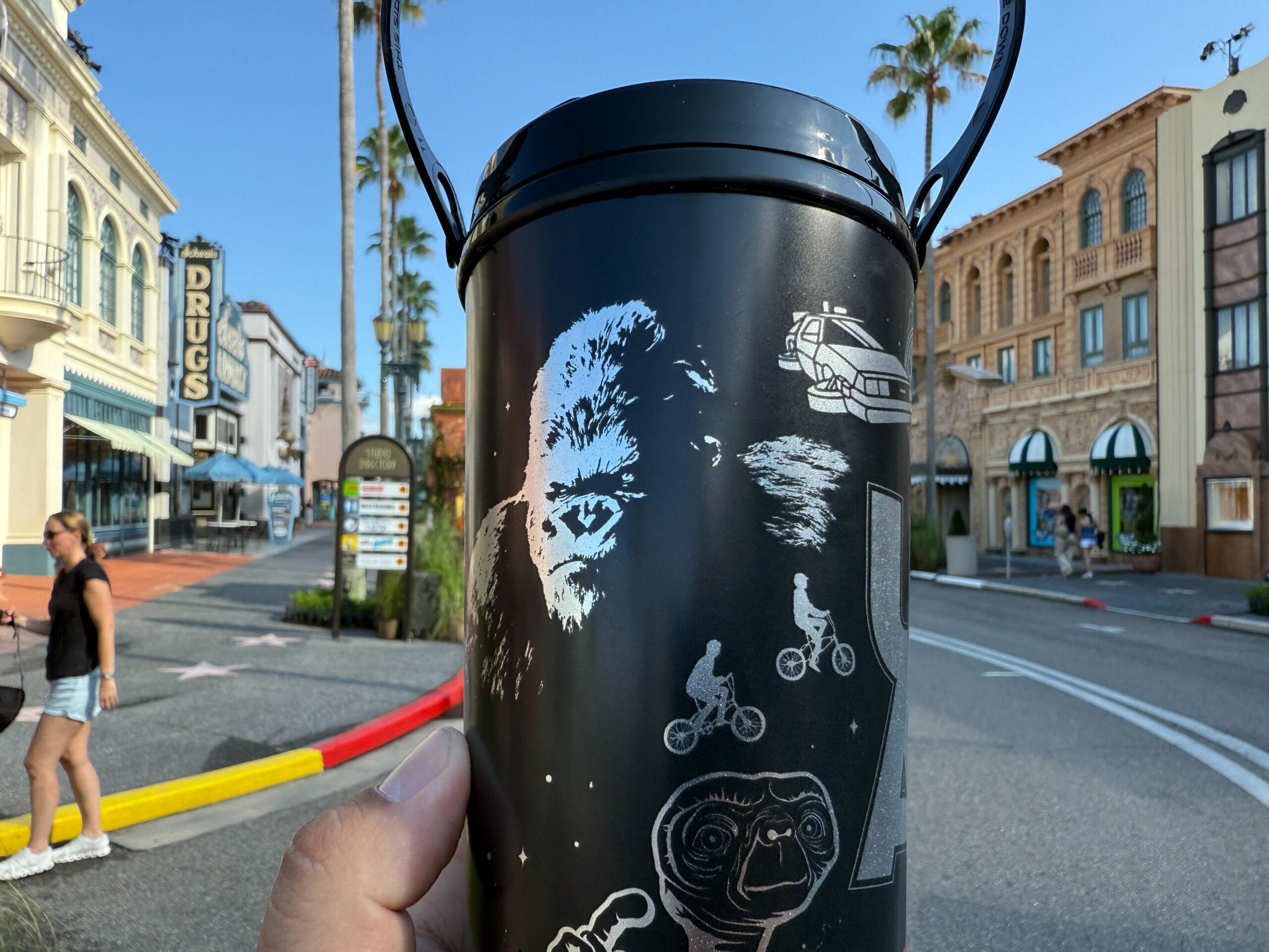 A hand holds a UOAP Coca-Cola Freestyle Cup with movie art on a sunny street lined with palm trees and vintage buildings.
