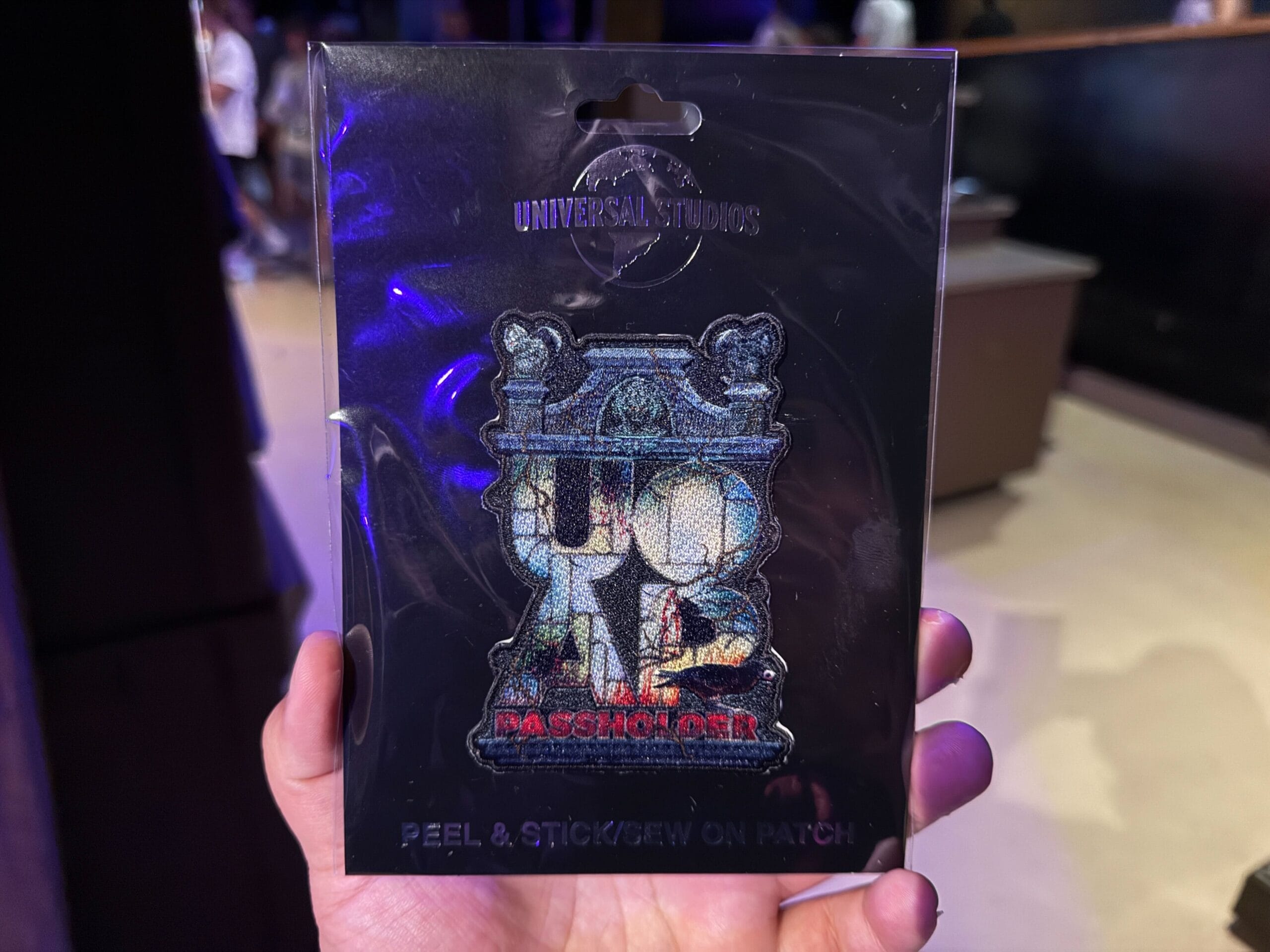 A hand holds a Universal Studios passholder patch for Halloween Horror Nights 34 in packaging inside a dimly lit room.