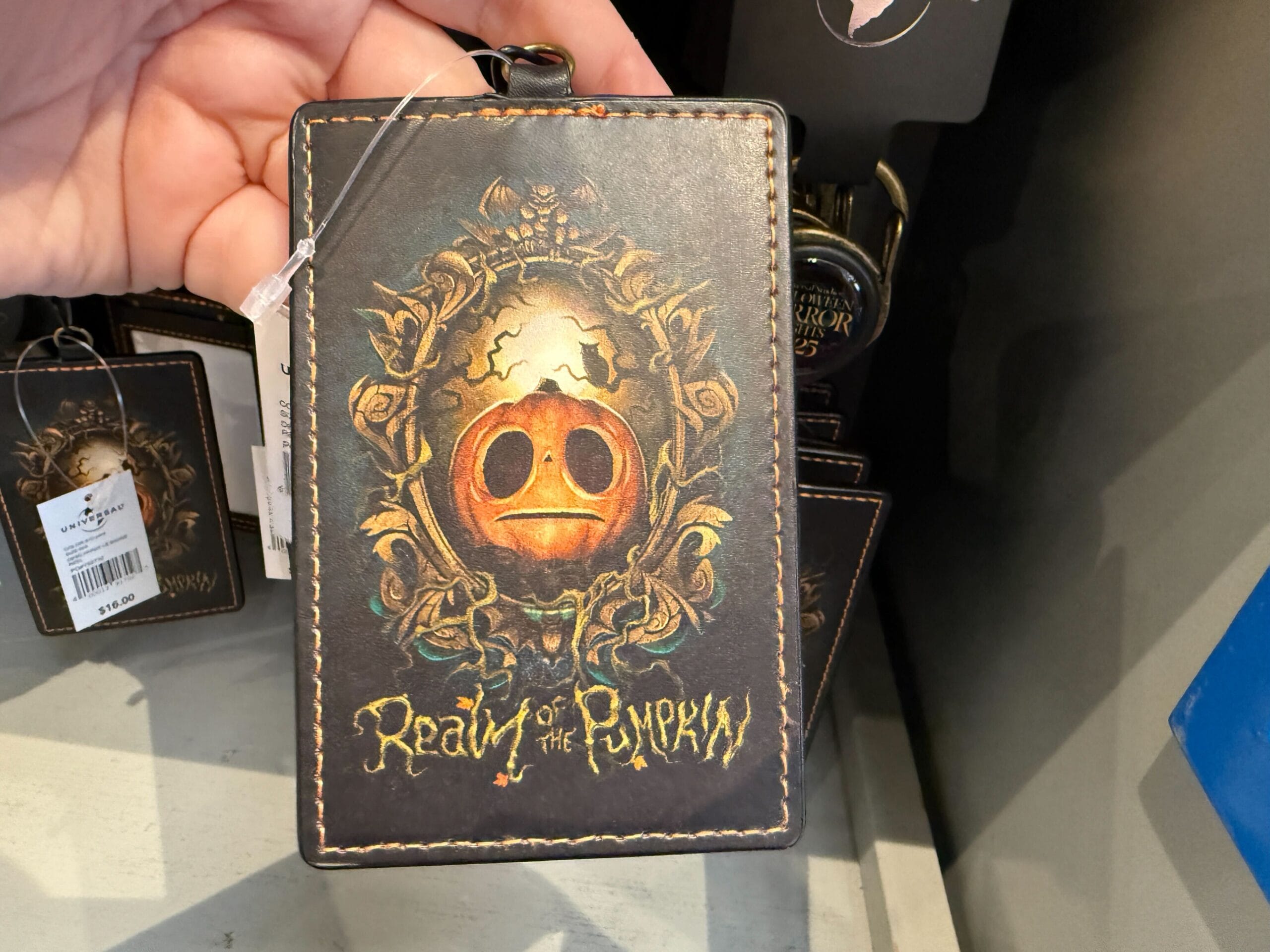 A hand holds a "Realm of the Pumpkin" card holder—an ornate piece of Halloween Horror Nights Merchandise.