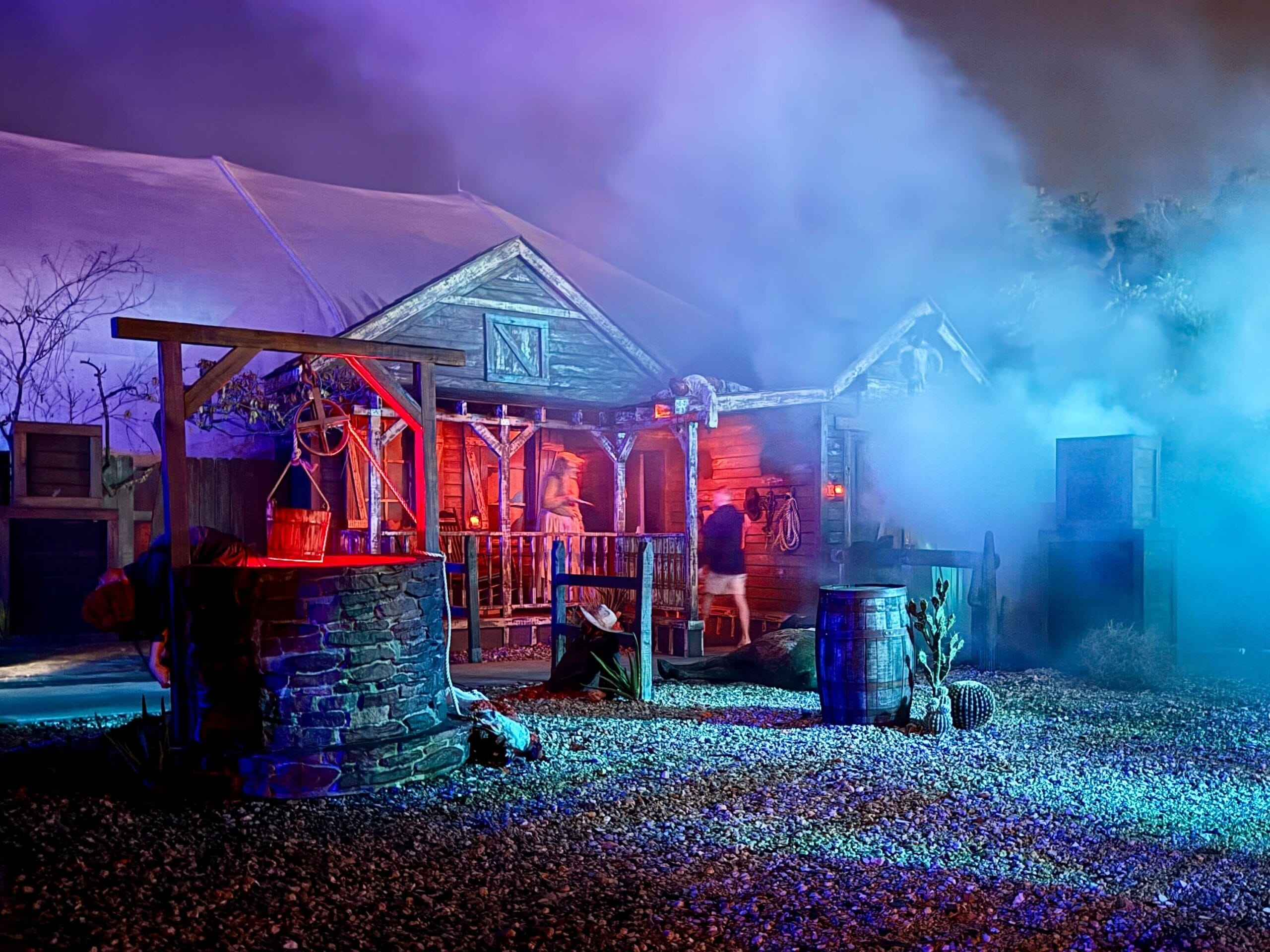 A foggy, colorfully lit old western house with barrels, a wooden well, and Demon Bounty Hunters at night.