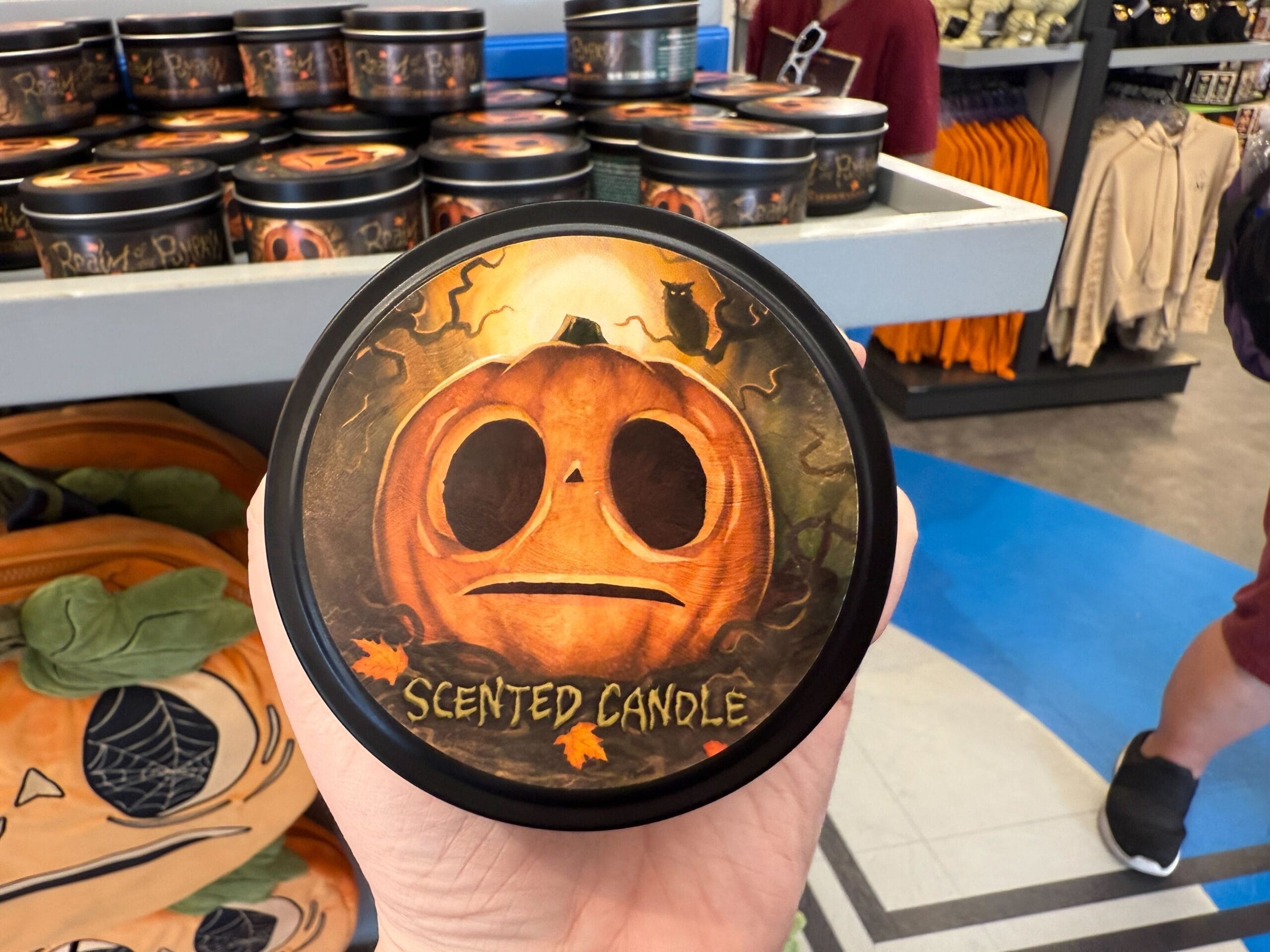 A hand holds a round tin labeled "Scented Candle" with a sad jack-o'-lantern face from Lil Boo Merchandise in a store display.