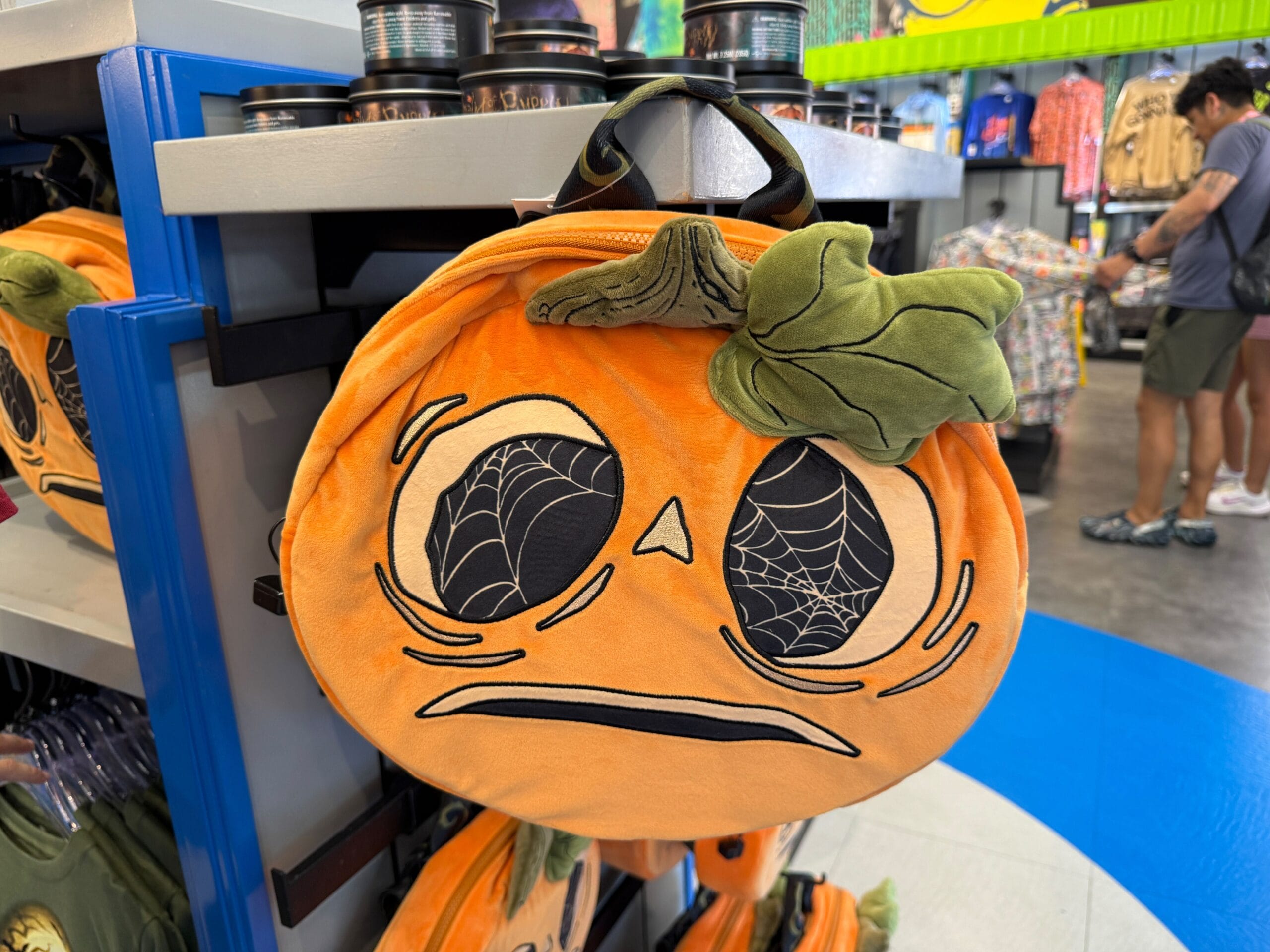 Plush pumpkin-shaped bag with sad eyes and spiderweb details from the Lil Boo Merchandise line on display shelf.