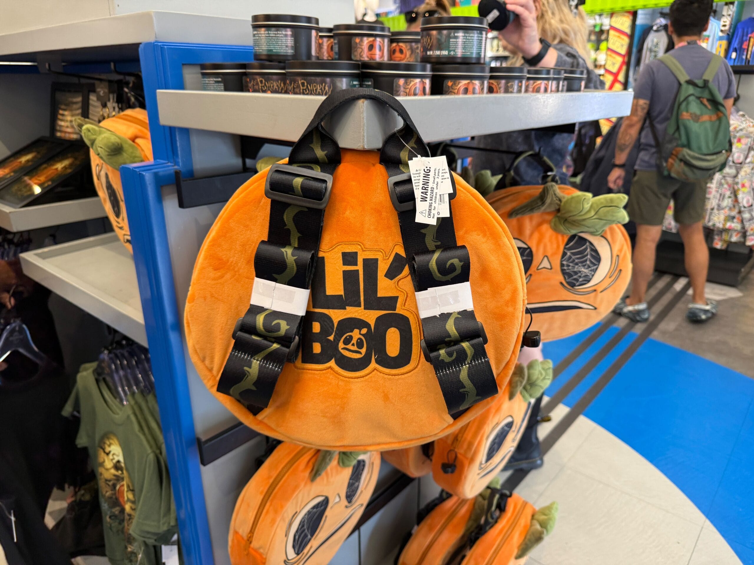 Round plush orange pumpkin bag with "Lil' Boo" text—adorable Lil Boo Merchandise with black straps, on store display.
