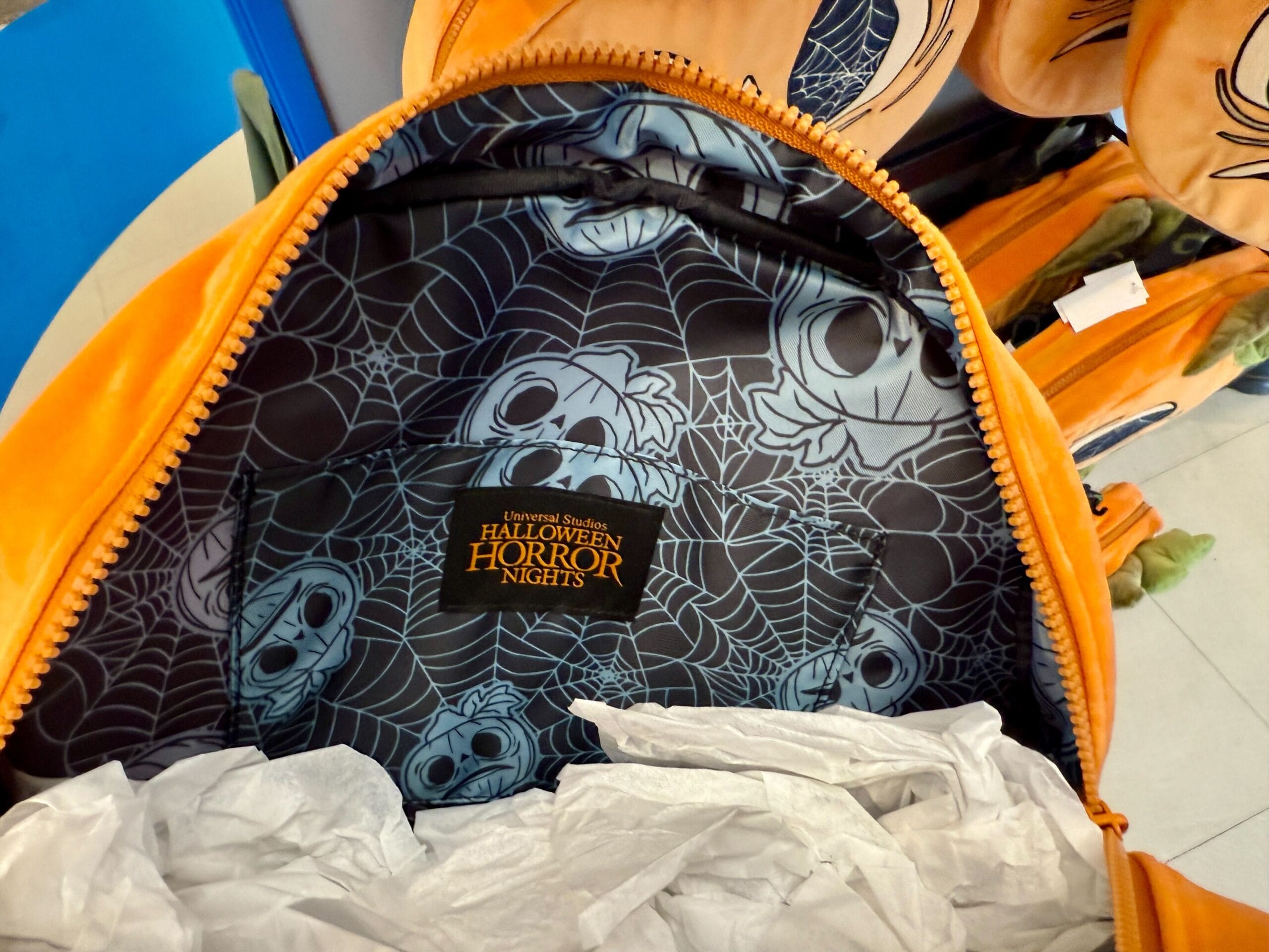 Open orange backpack with "Halloween Horror Nights" label, spiderweb lining, and ghost faces—perfect for Lil Boo merchandise fans.