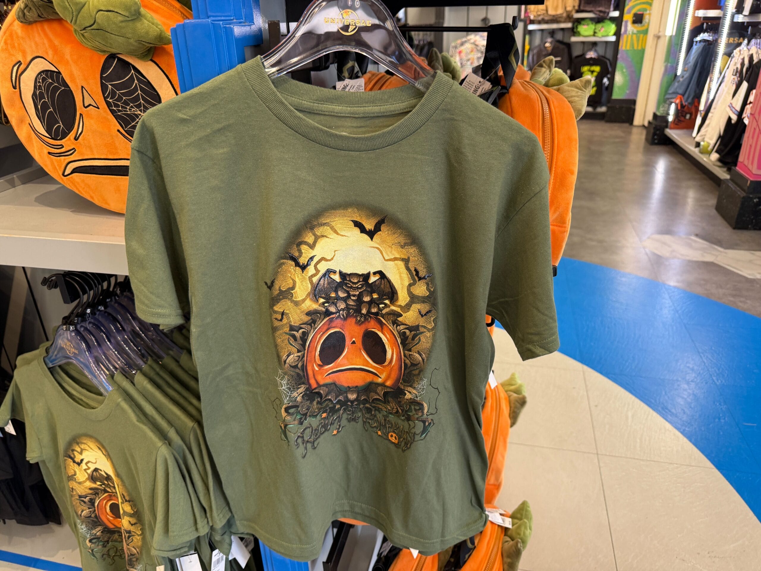 Green T-shirt from Lil Boo Merchandise with a sad pumpkin face and bat design, displayed near pumpkin plushies.