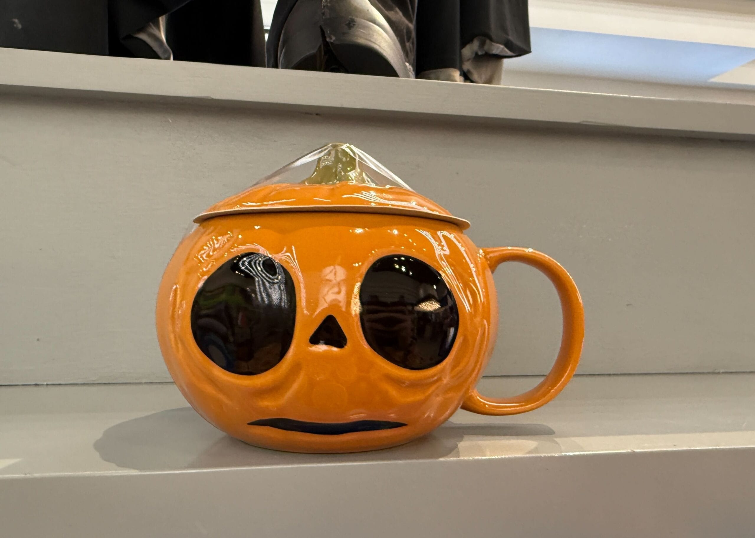 Orange pumpkin-shaped Lil Boo Merchandise mug with a lid, black eyes, and a sad mouth, sitting on gray steps.