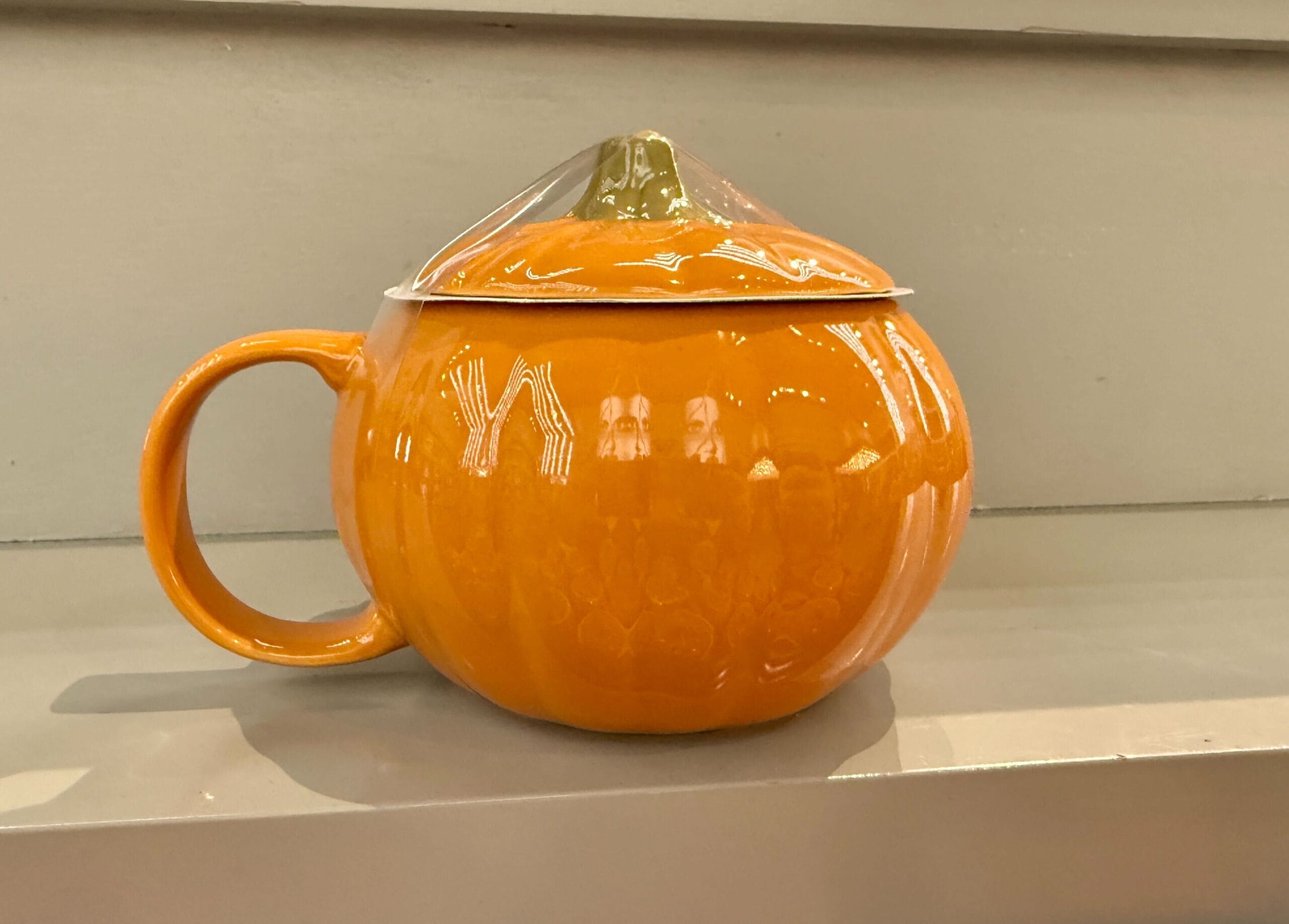 Orange pumpkin-shaped mug with lid from the Lil Boo Merchandise line, displayed on a light-colored shelf.