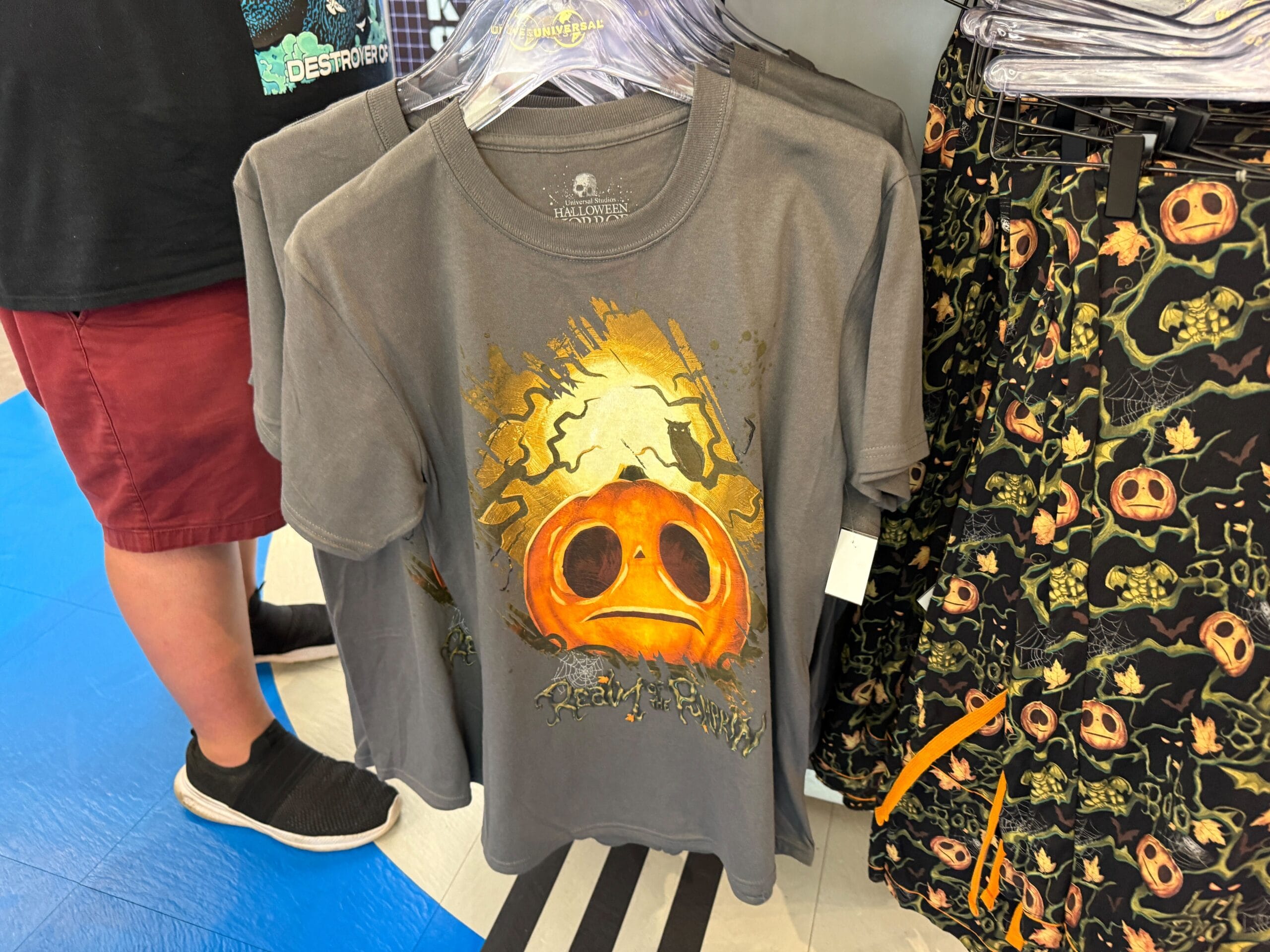 Gray t-shirt with a cracked pumpkin face and "Beauty Fright" text, part of Lil Boo Merchandise, displayed on a rack.