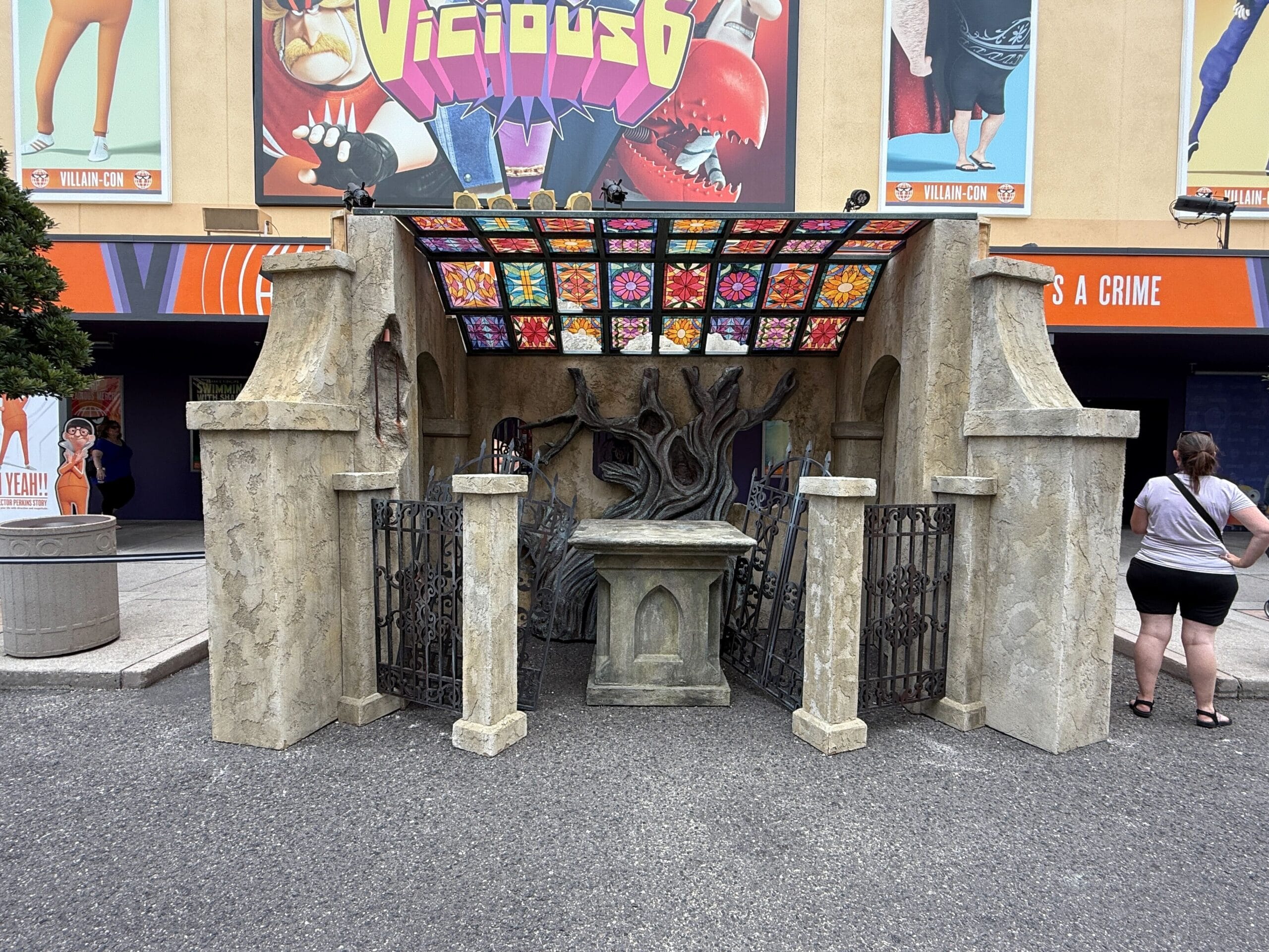 A stone altar with a twisted tree and stained-glass roof stands outdoors near HHN 34 construction murals.