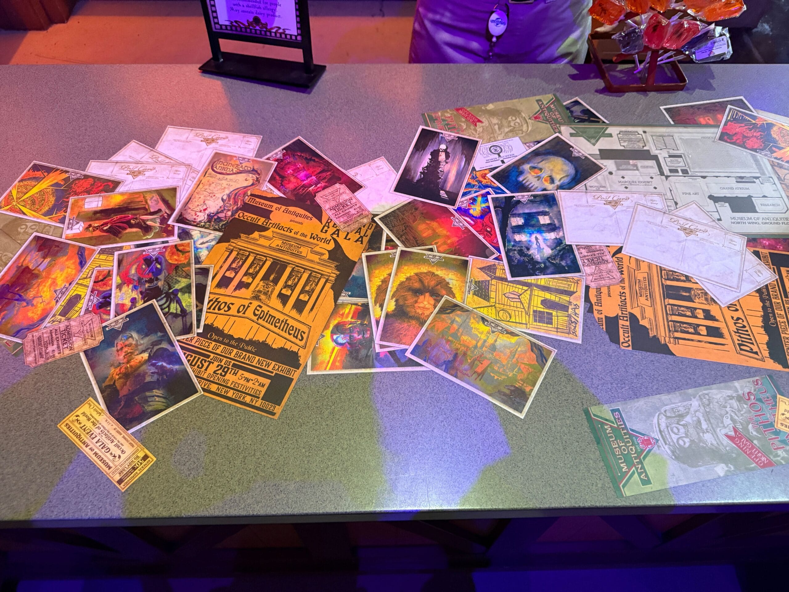 A tabletop covered with colorful postcards, tickets, and maps in a dimly lit, purple-toned setting.
