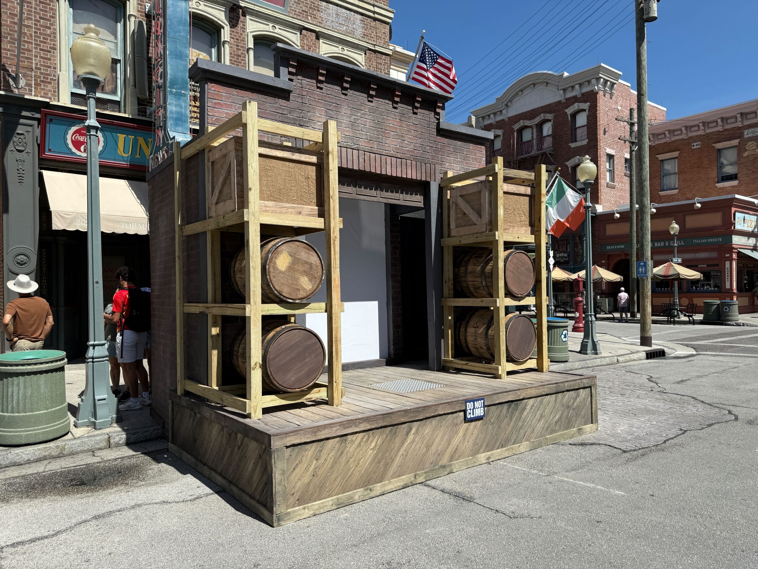 A stage with stacked wooden barrels on a city street, next to brick buildings and flags under a clear blue sky.