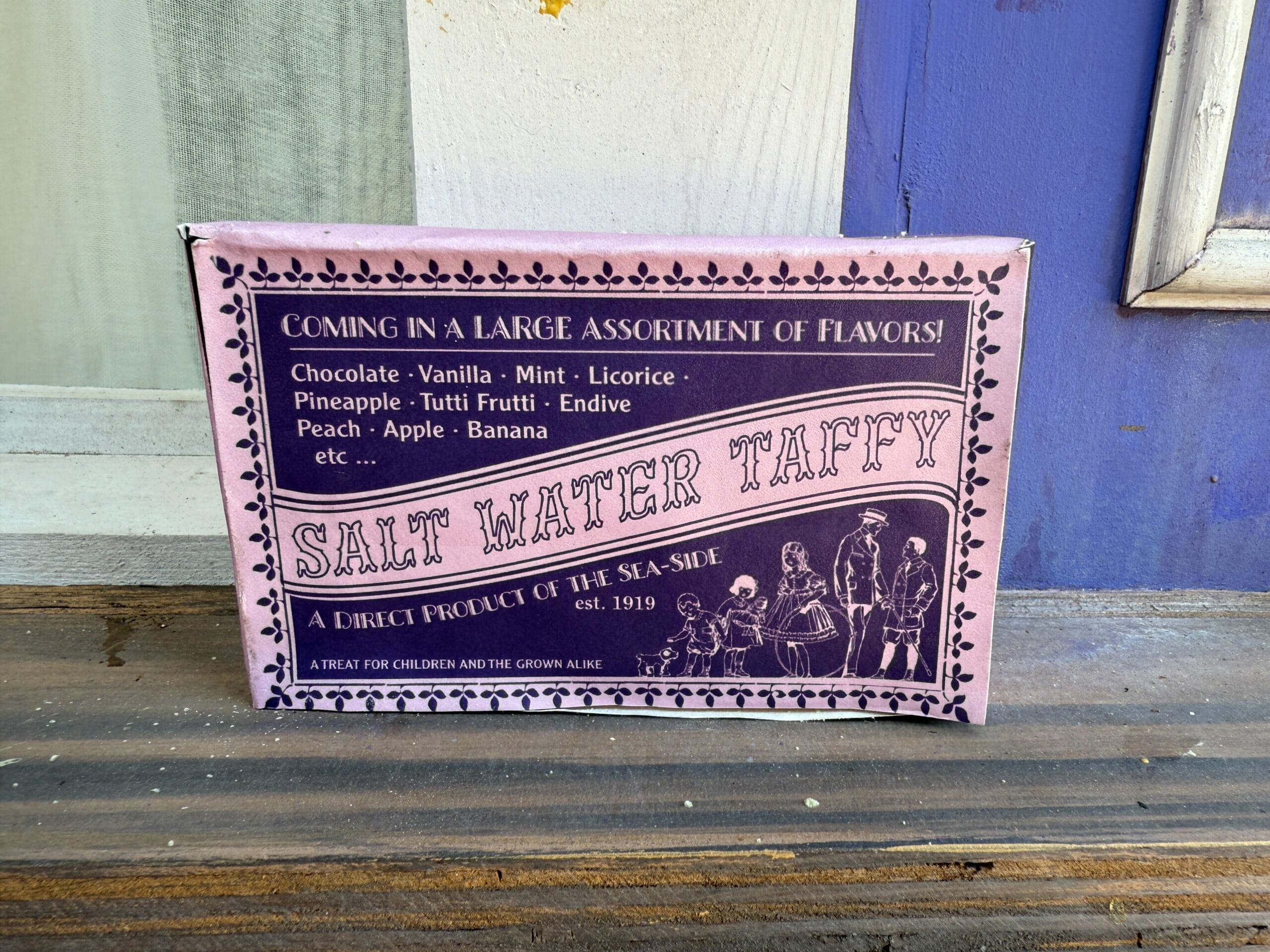 A pink box of salt water taffy lists flavors like chocolate, mint, and banana on a wooden ledge near a purple wall.