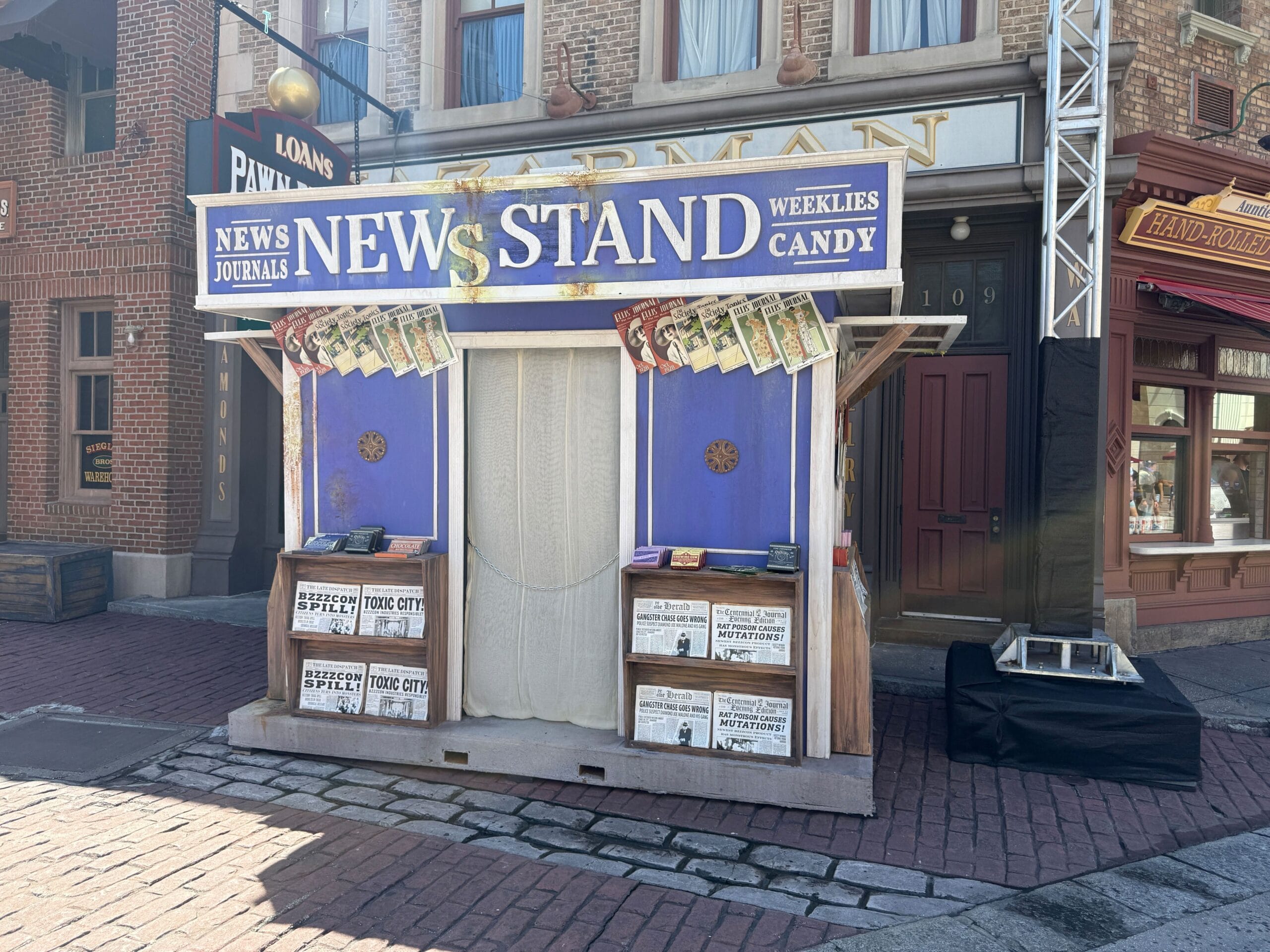 A vintage newsstand with newspapers and candy, set on a cobblestone street in front of brick buildings.