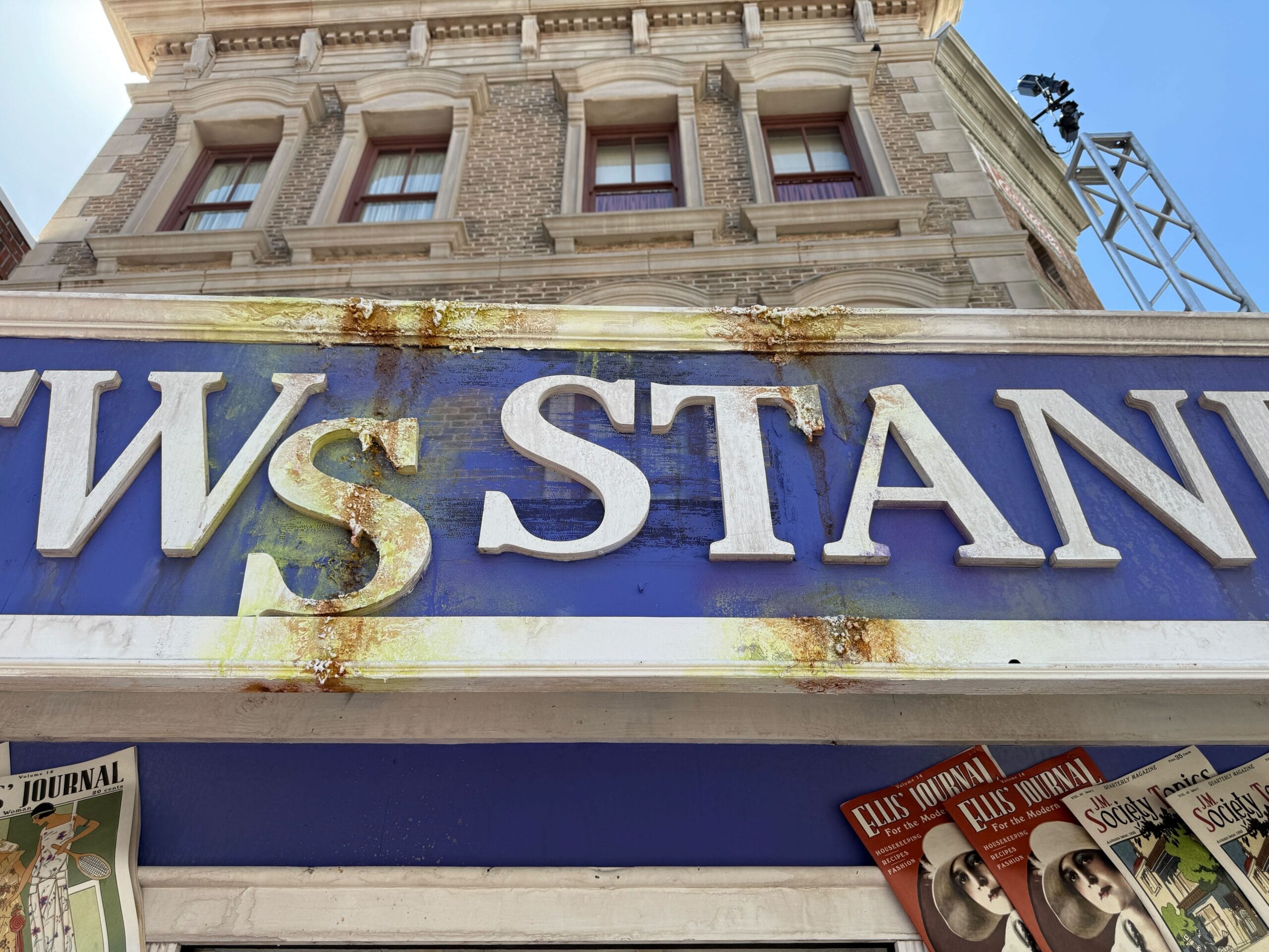 Close-up of a weathered blue newsstand sign with rust stains and vintage magazines displayed below.