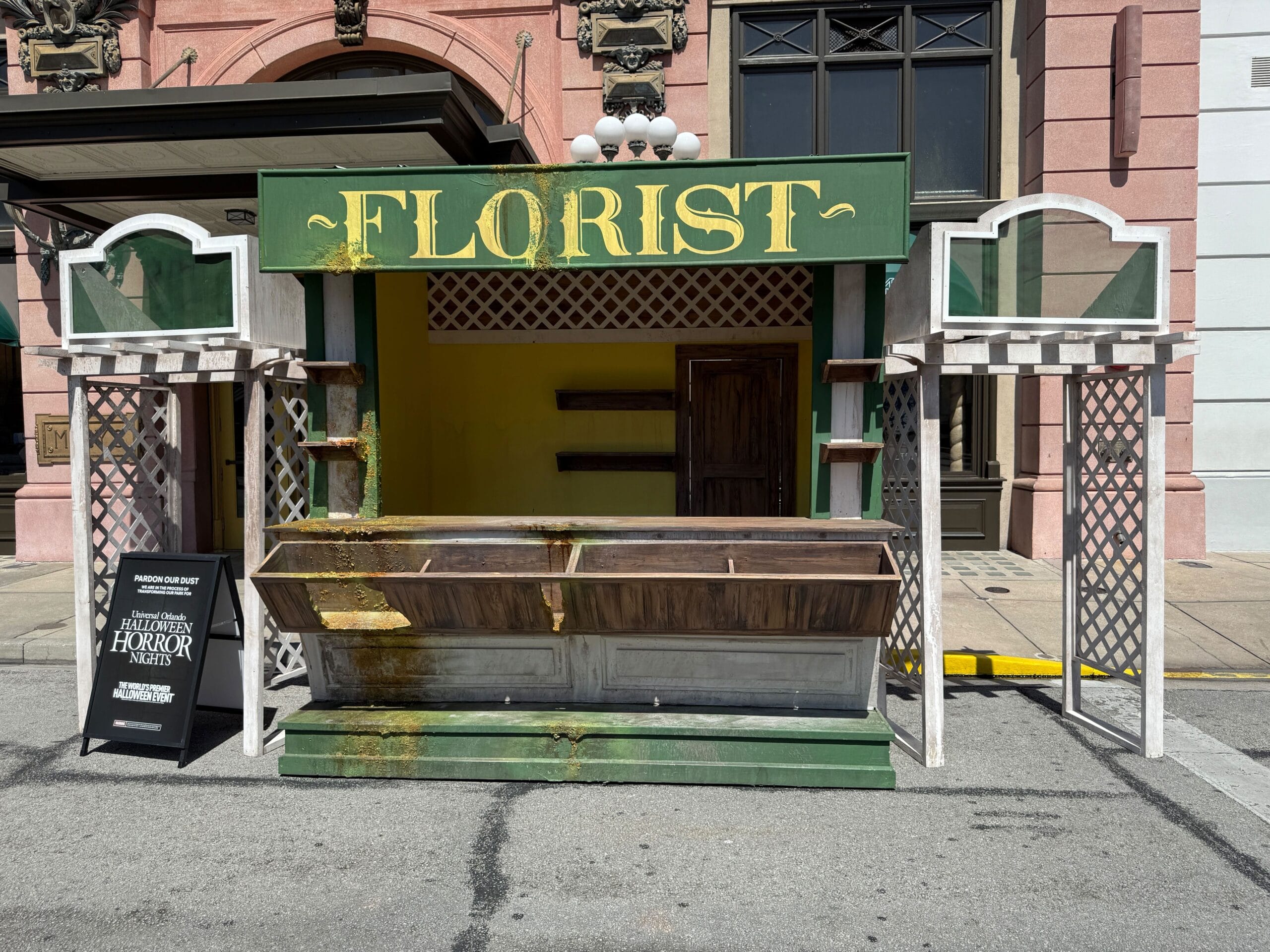 An empty, weathered florist stand with a green sign and a small black event sign, set on a city street.