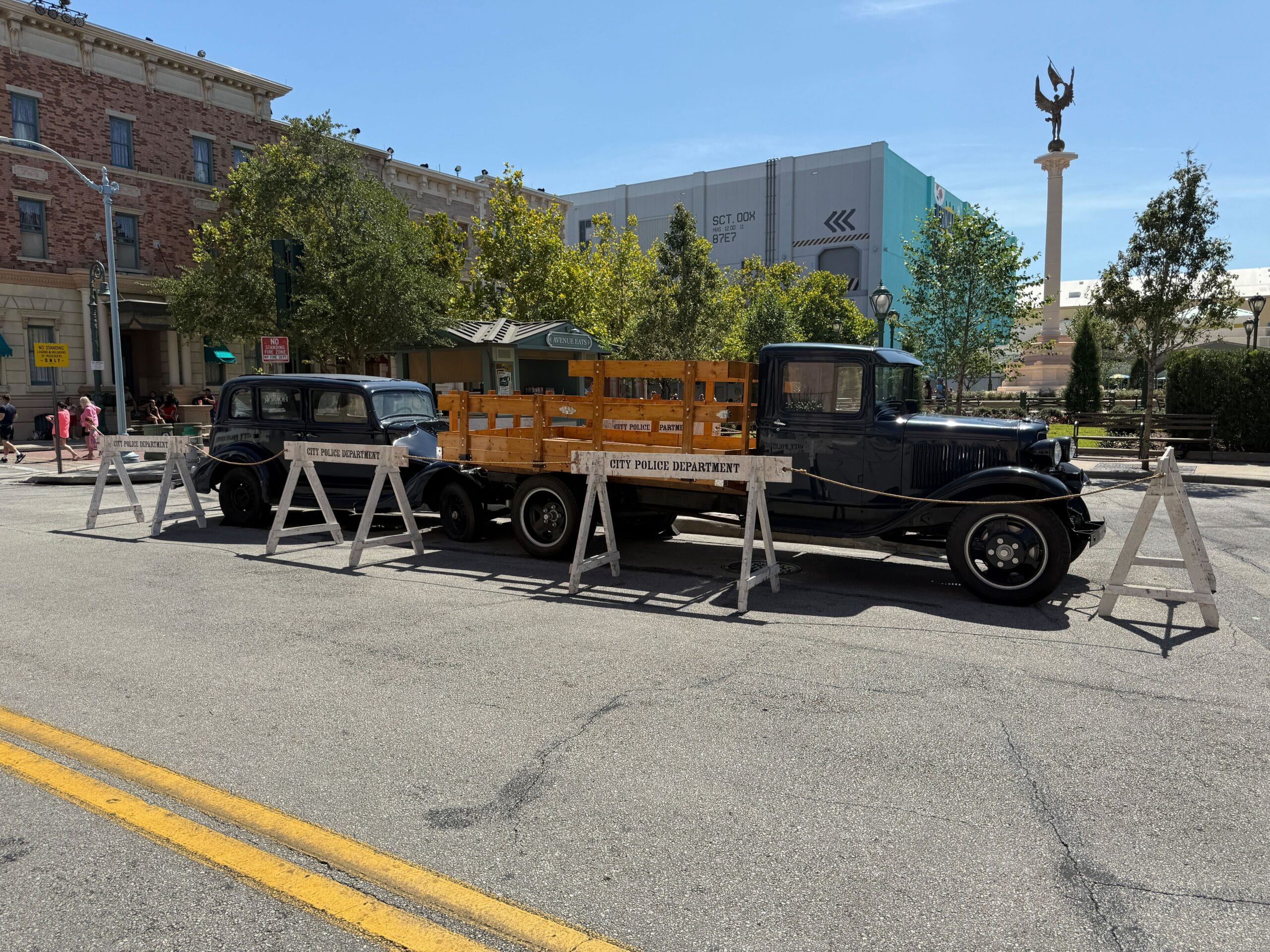Two vintage vehicles behind wooden barricades on a city street, with buildings and trees in the background.