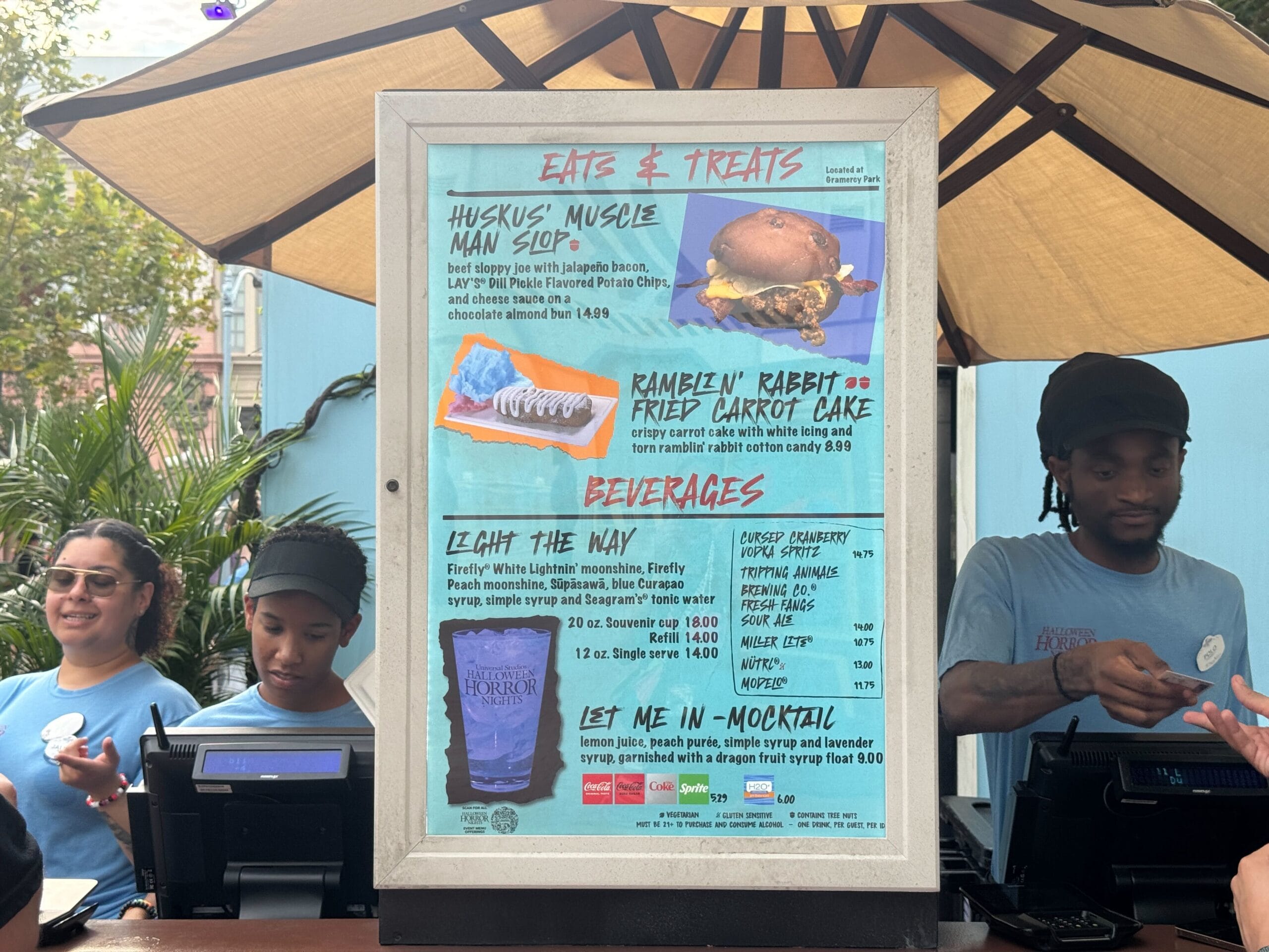 Menu board for eats and drinks with staff standing behind registers at an outdoor food stand.