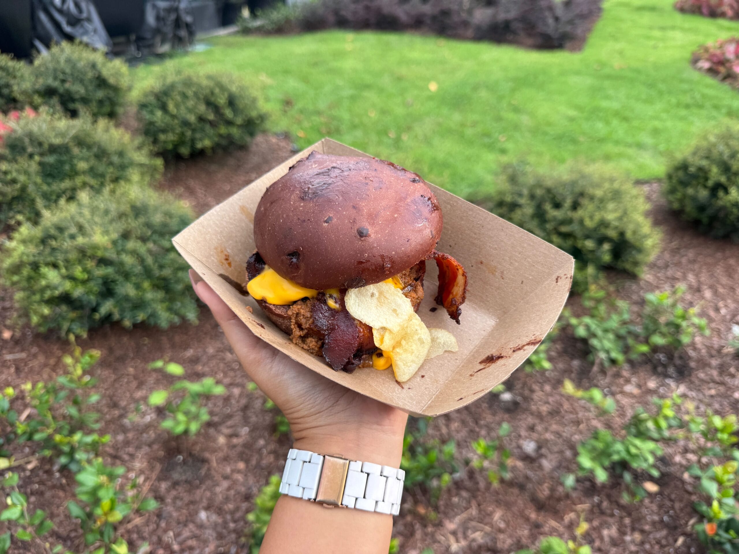 A hand holds a burger with cheese, bacon, and chips in a paper tray outdoors, with green bushes in the background.
