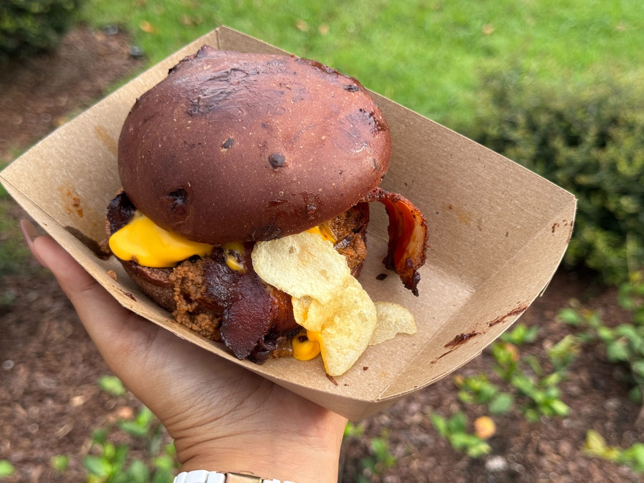 A hand holds a burger with bacon, cheese, and potato chips in a cardboard tray, outdoors with greenery in the background.