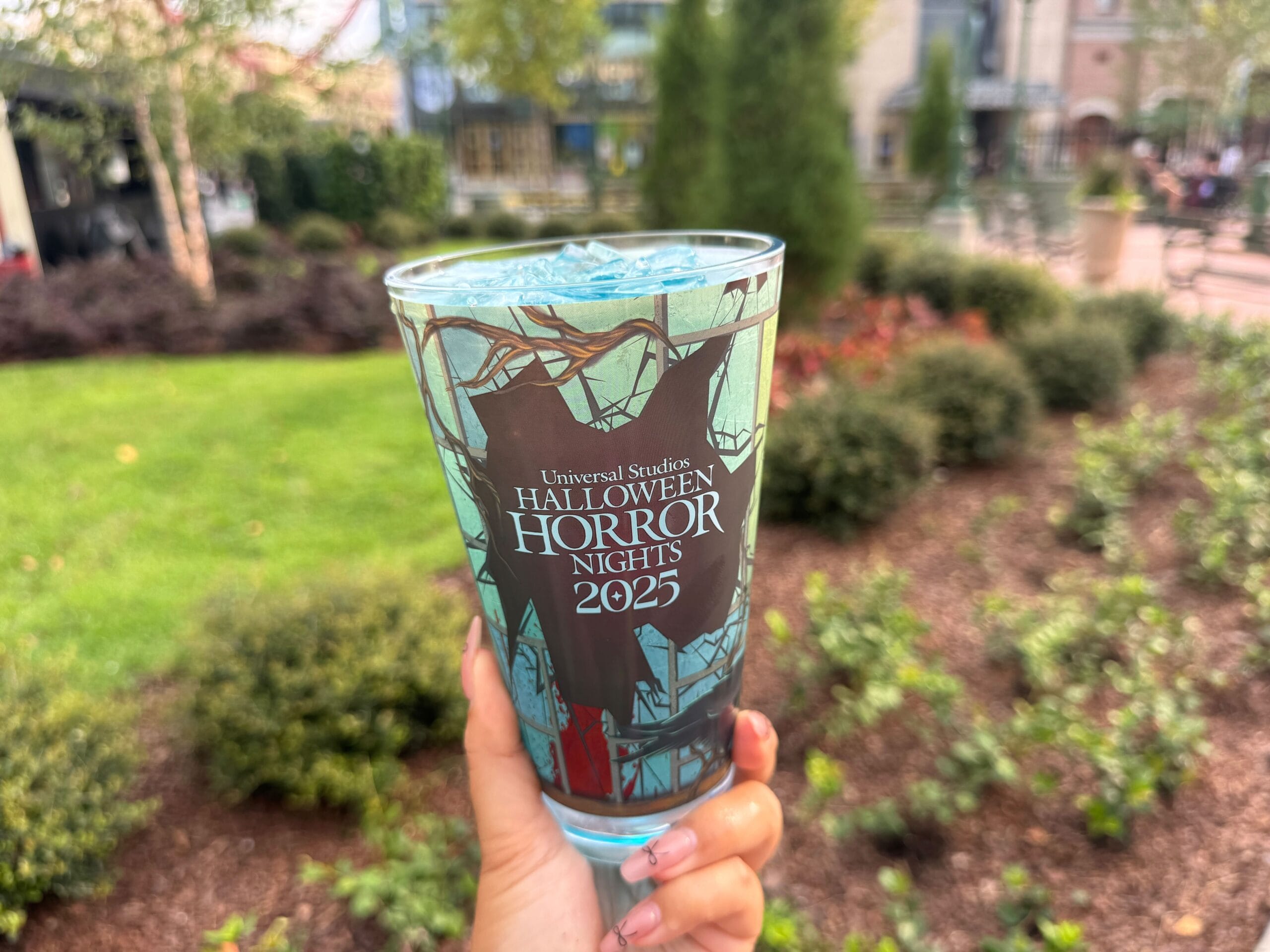 A hand holds a blue drink in a "Halloween Horror Nights 2023" cup at Universal Studios, with greenery in the background.