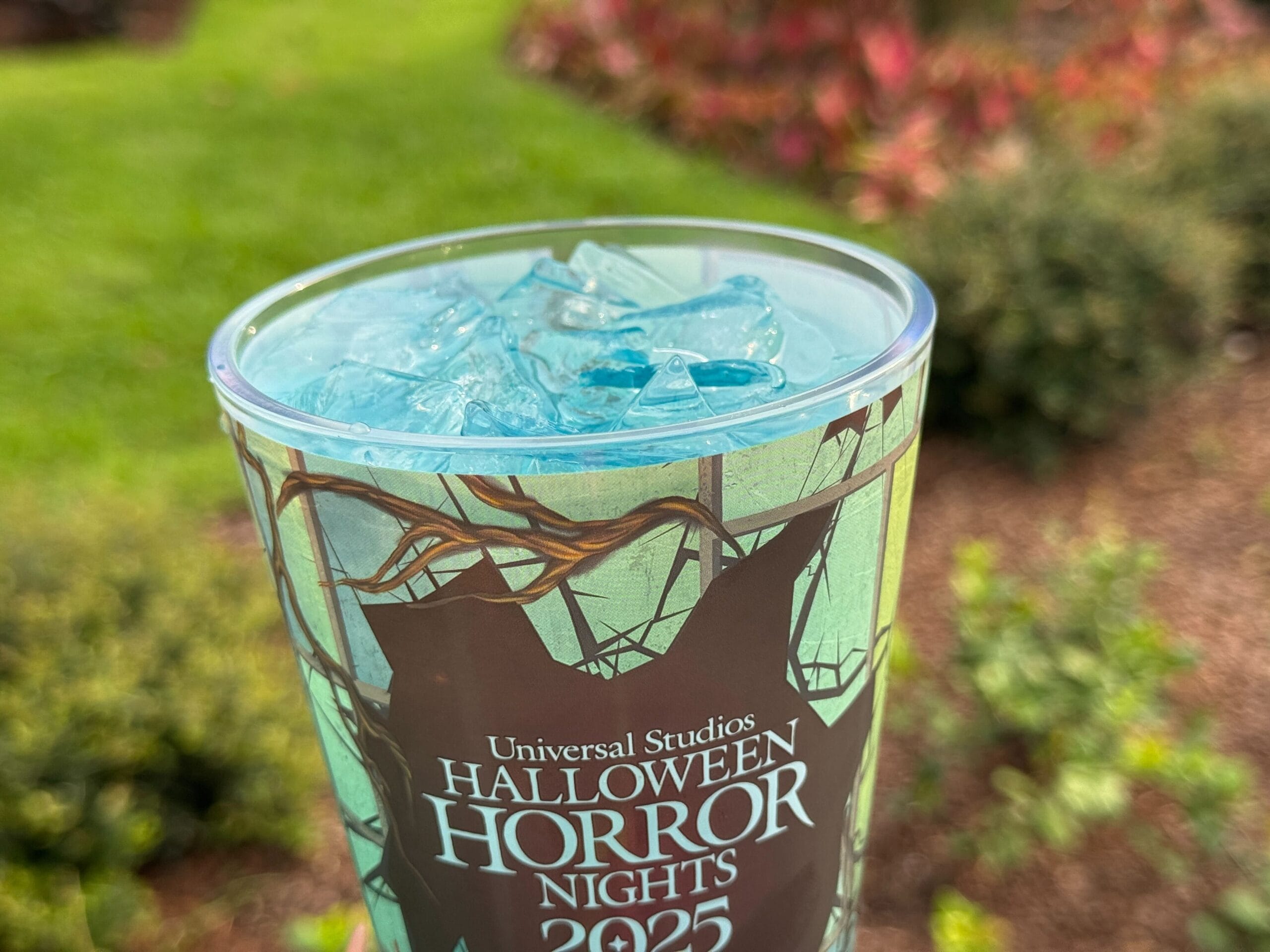 A Halloween Horror Nights 2025 cup filled with blue ice drink, with greenery and plants blurred in the background.