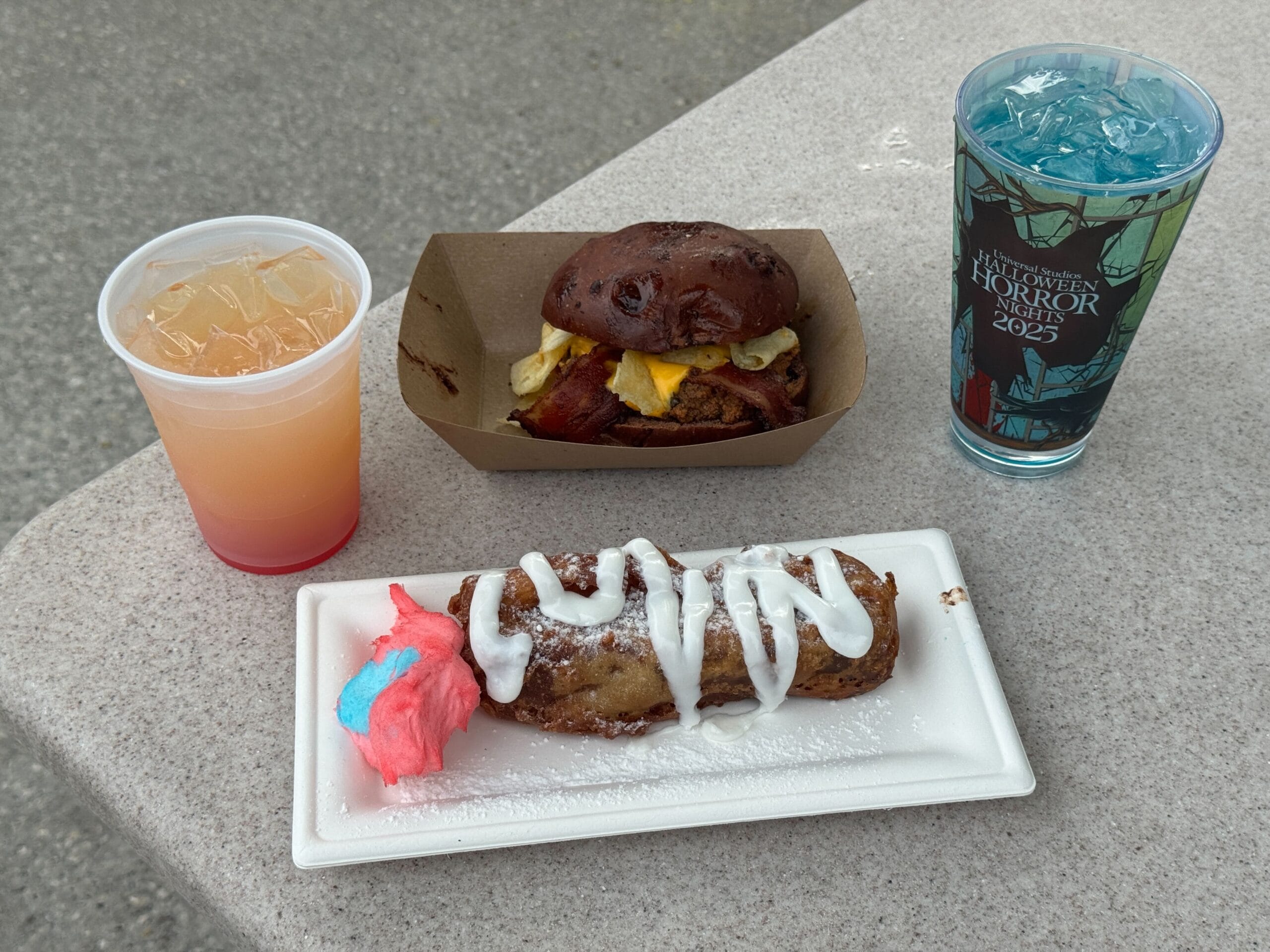 Two colorful drinks, a burger with cheese in a pretzel bun, and a dessert pastry with icing and pink-blue cotton candy.