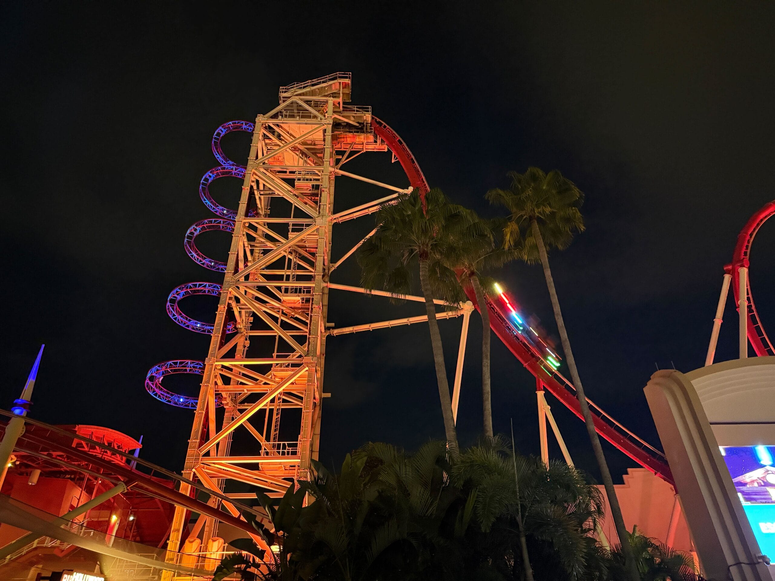A tall, illuminated Hollywood Rip Ride Rockit coaster twists at night, framed by palm trees and a dark sky.