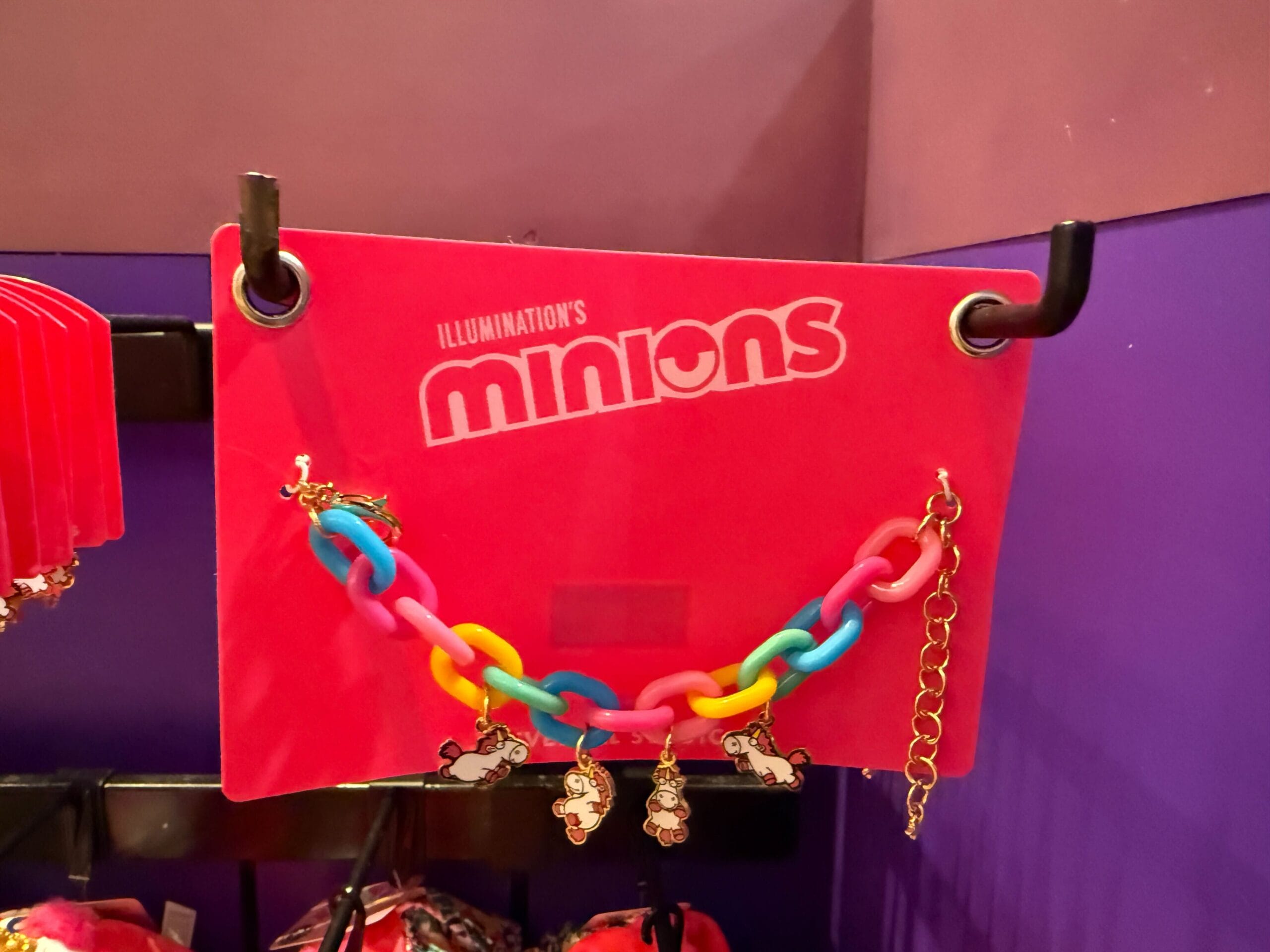 A colorful Minions bracelet with Fluffy Unicorn charms hangs on a display card against a purple wall.