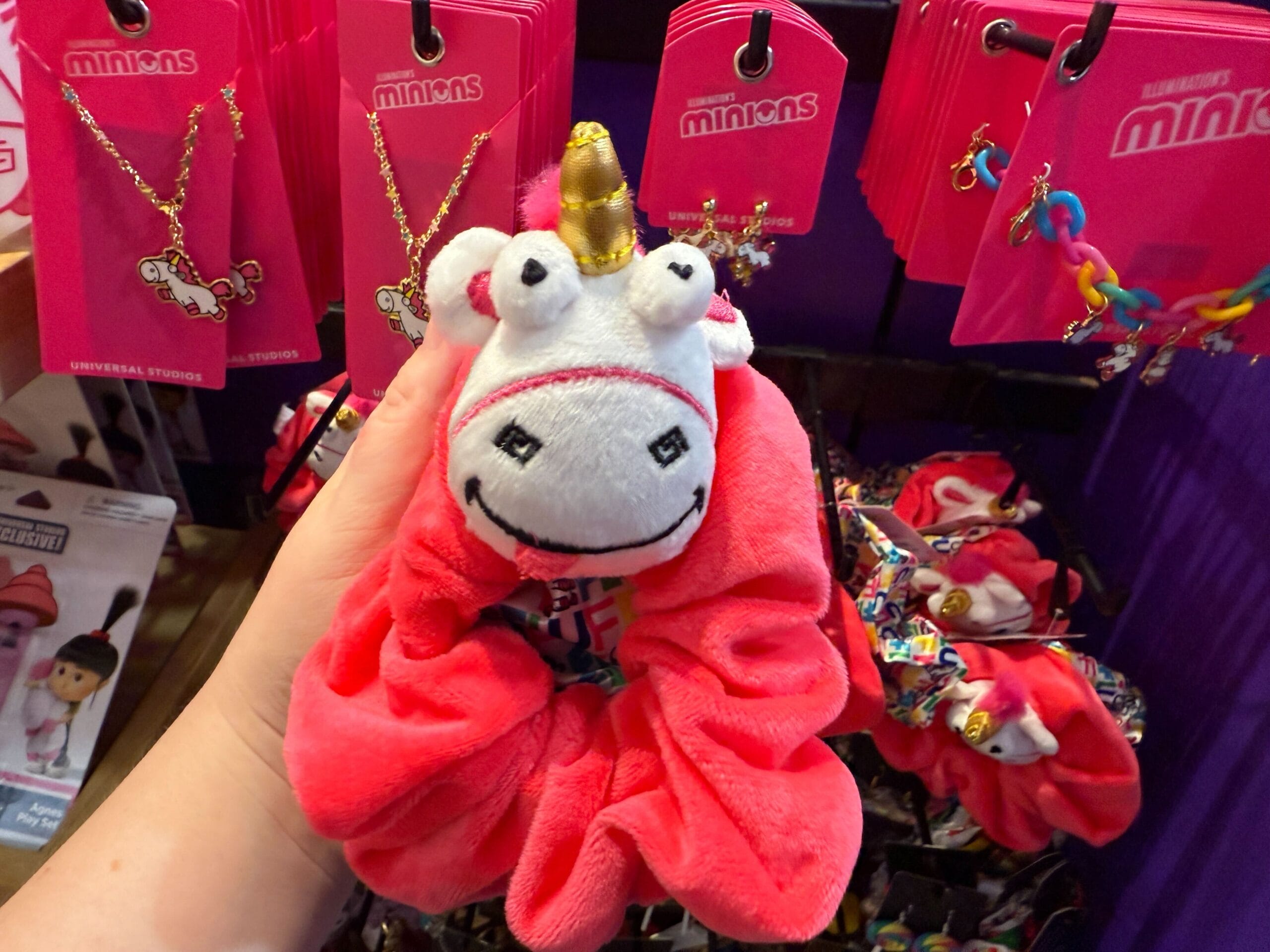 A hand holds a pink Fluffy Unicorn scrunchie near Minions merchandise at Universal Orlando Resort.