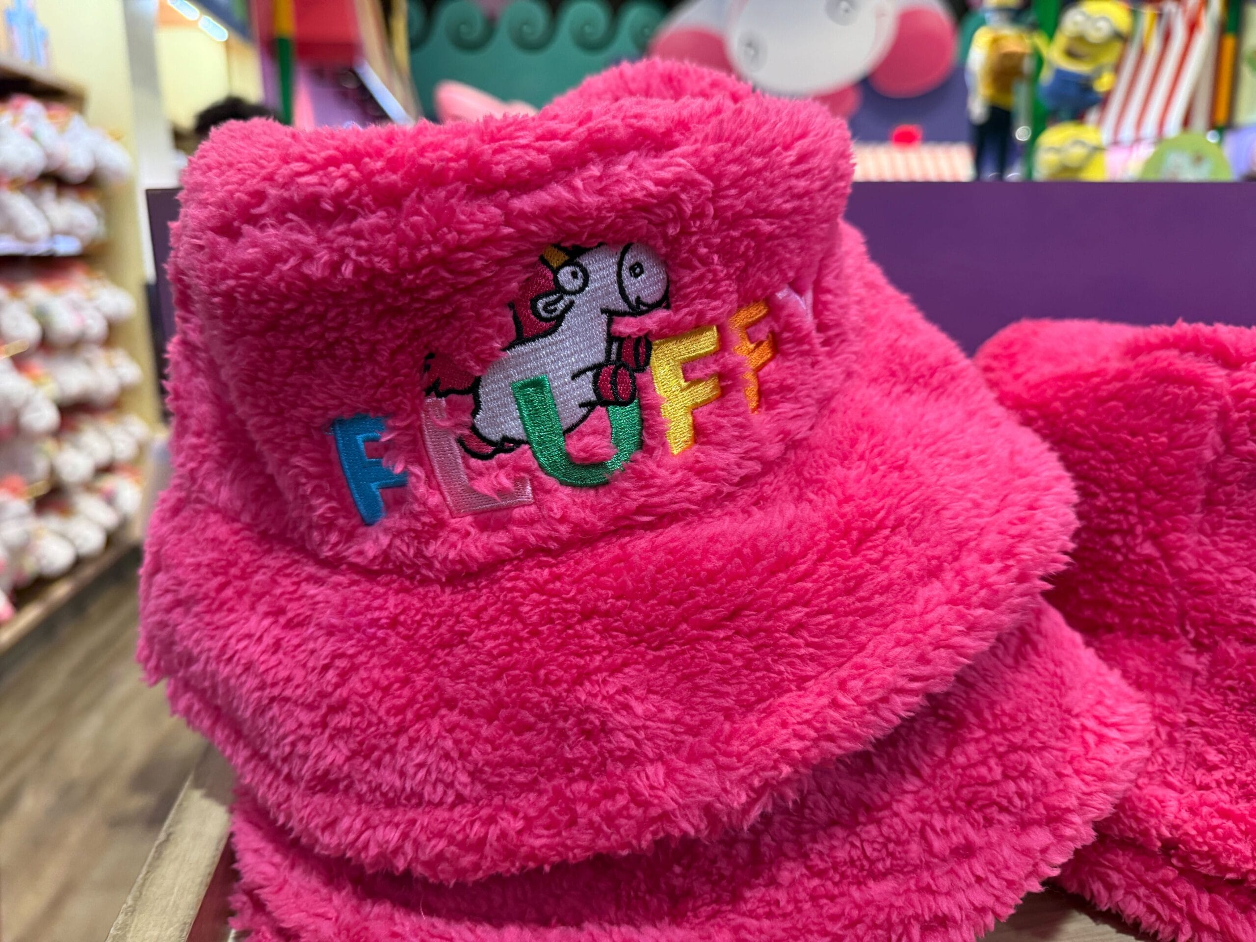 A bright pink fuzzy hat with "FLUFFY" and a Minions Fluffy Unicorn cartoon embroidered on it.