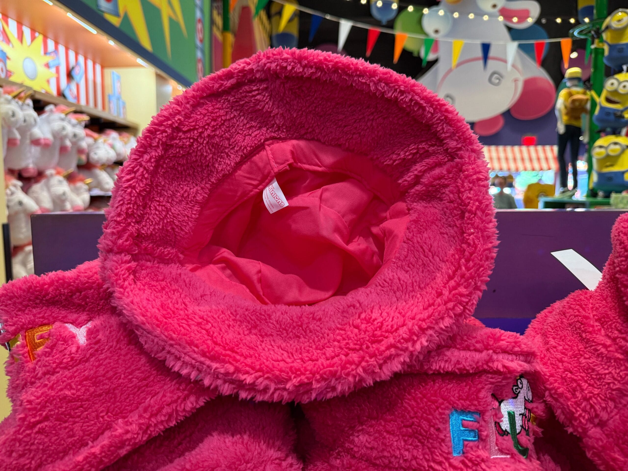 A fuzzy pink hooded jacket, inspired by the Fluffy Unicorn, on display in a colorful toy store with plush toys all around.