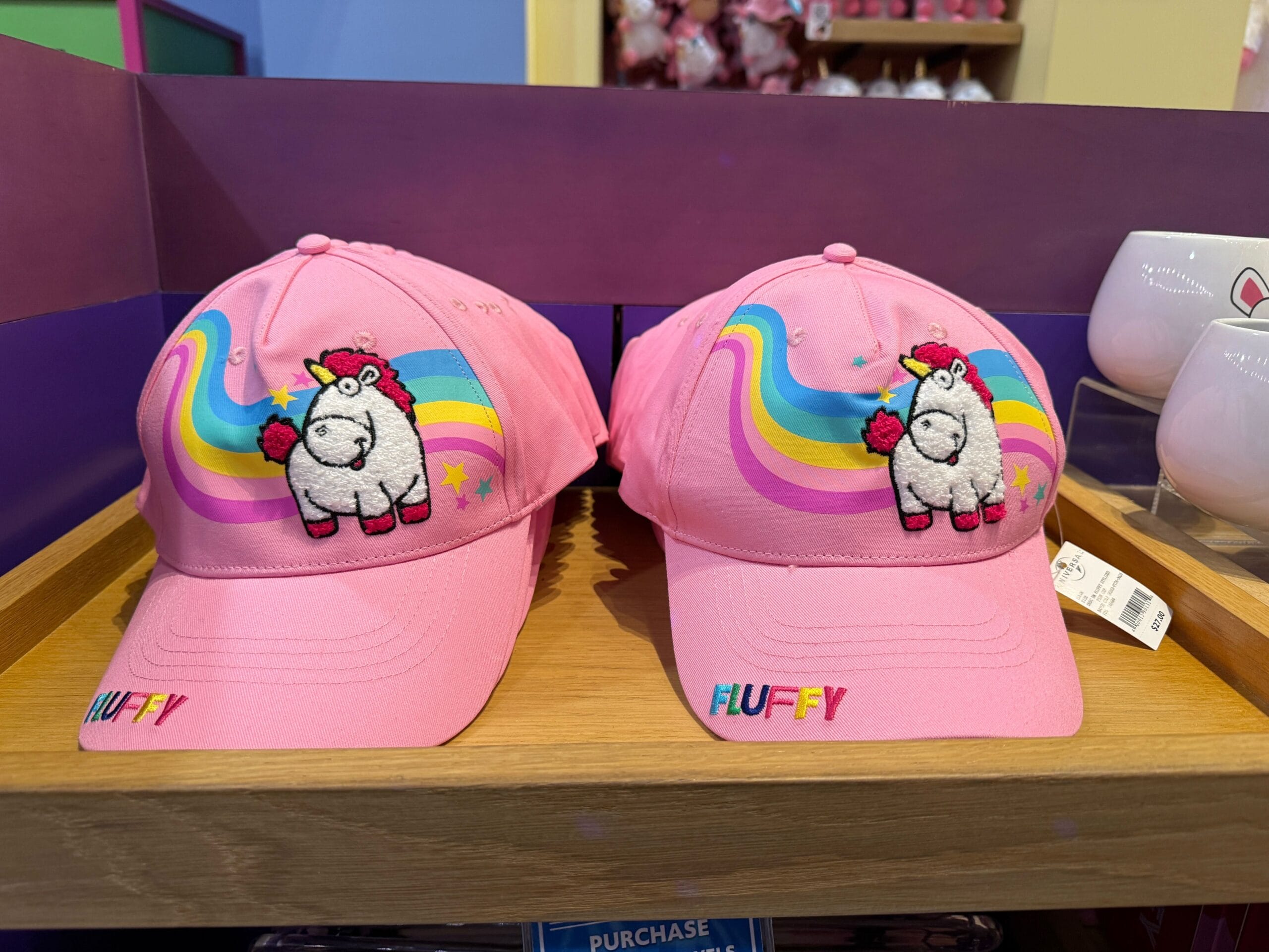 Two pink caps with a Fluffy Unicorn and rainbow design, plus "Fluffy" text, displayed on a wooden shelf.